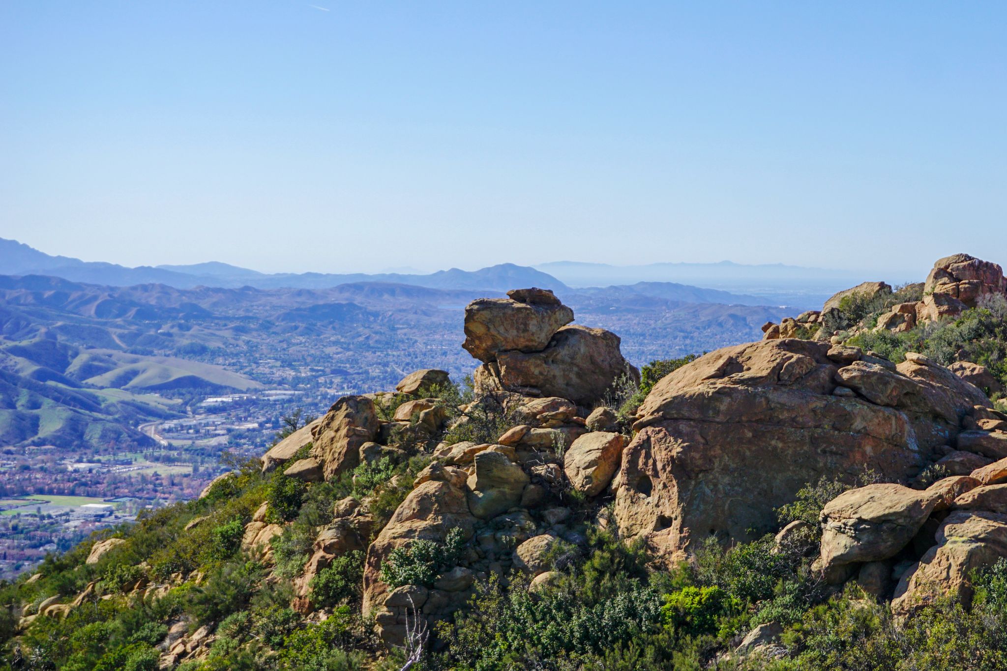 Large rock formations on a hillside overlooking a distant town and mountains. - Rocky Peak Trail – Rocky Peak Trail loop from Santa Susana Pass State Historic Park