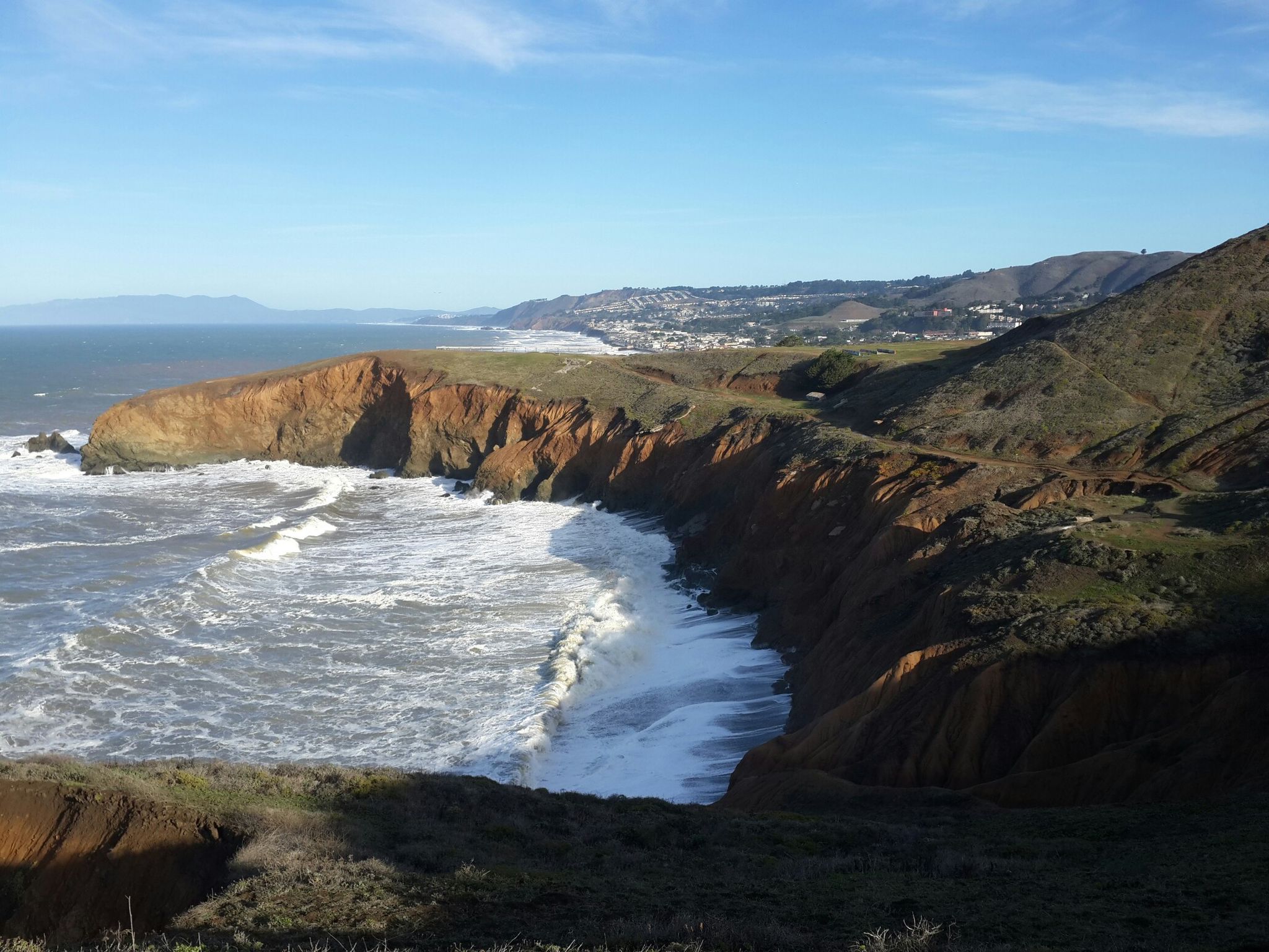 Dramatic reddish cliffs meet the ocean, with waves crashing below on a sunny day. - Rocky Shoreline at Mori Point – The Point loop from Cabrillo Highway & Westport Drive