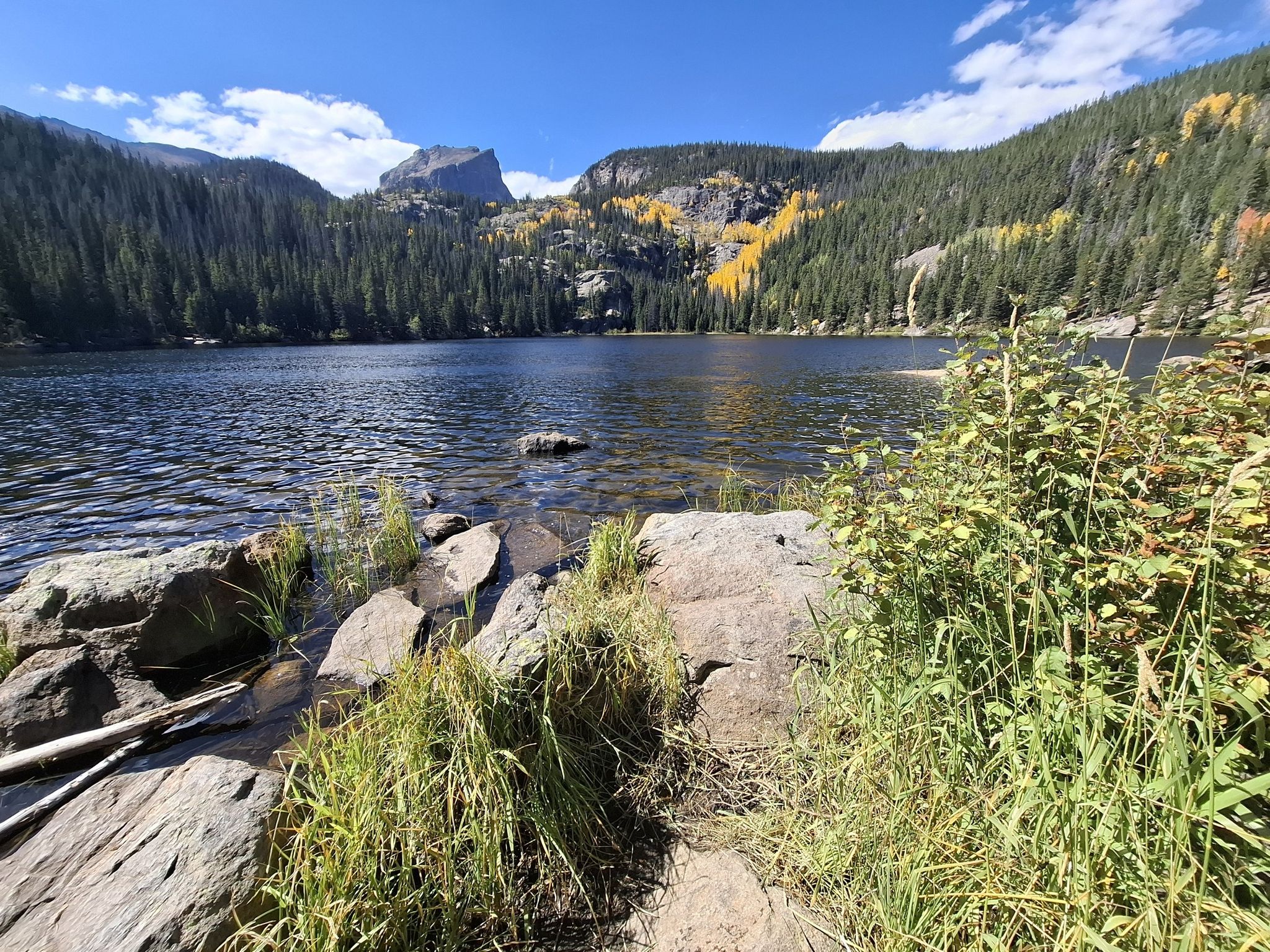 Clear lake with rocky shore, pine forests, and distant mountains under blue sky. - Flattop Mountain Trail