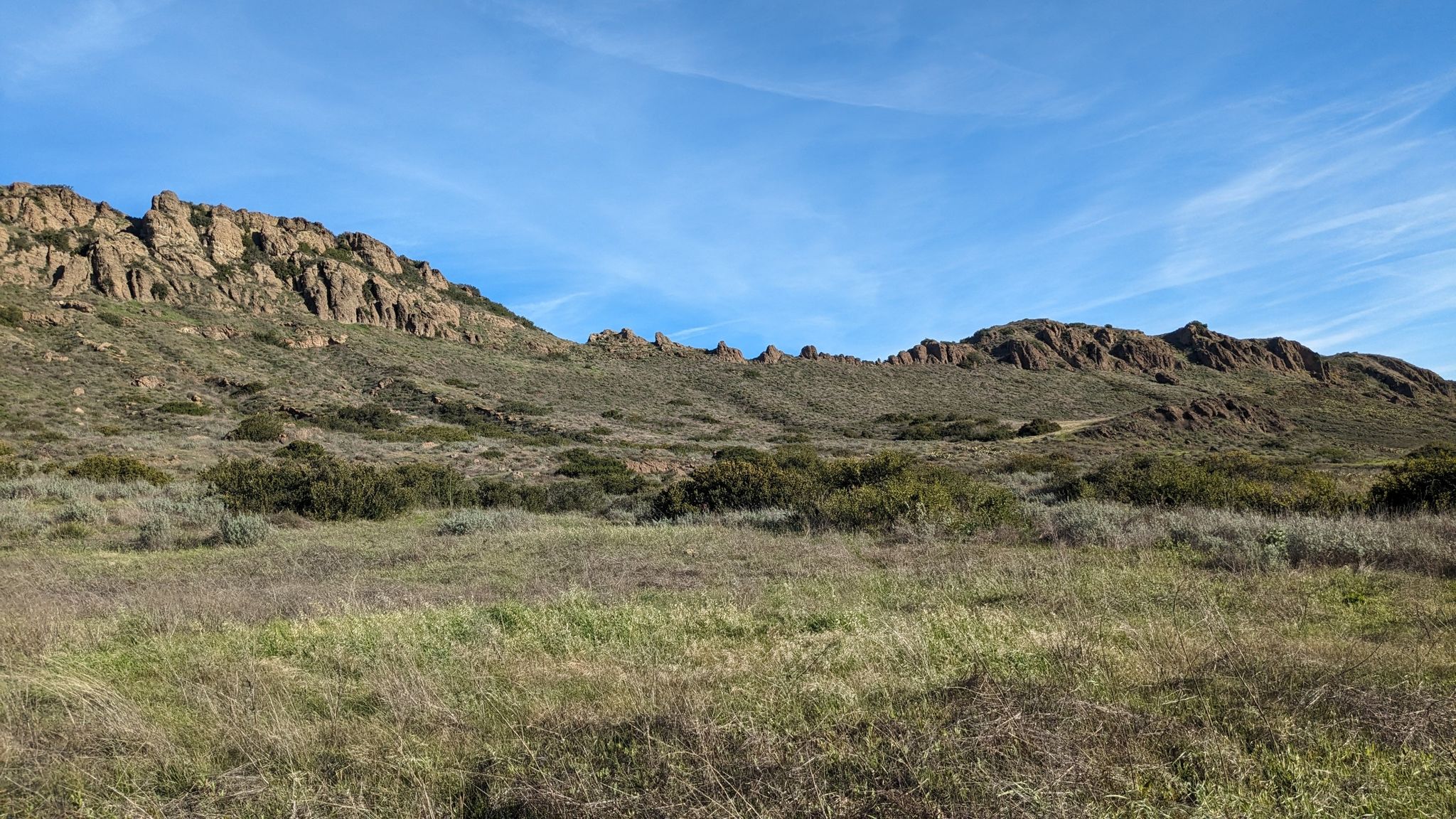 Wide grassy field with rocky hills and mountains in the background under a blue sky. - Stagecoach Bluff – North Tepee Trail loop from California Lutheran University