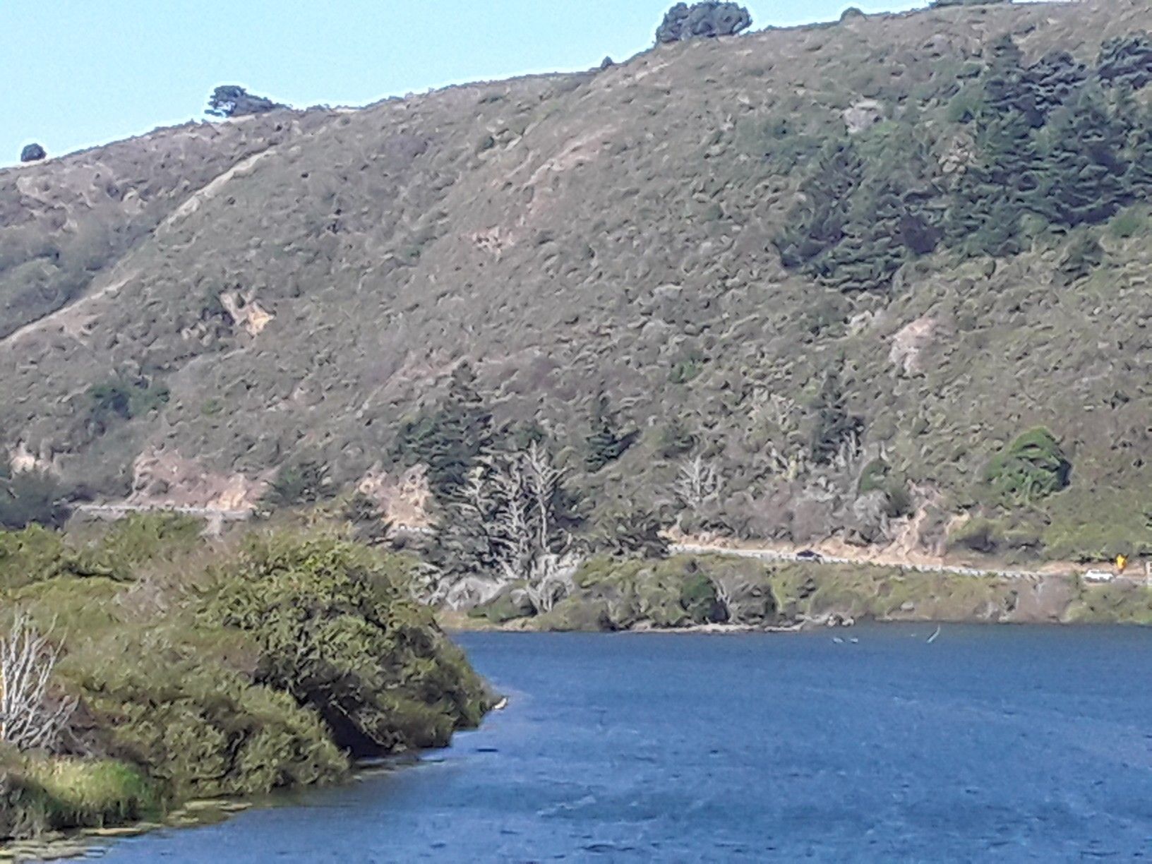 Blue water in foreground, green hills with a winding road under bright daylight. - Elk Creek Beach Overlook – Greenwood State Beach loop from Greenwood State Beach