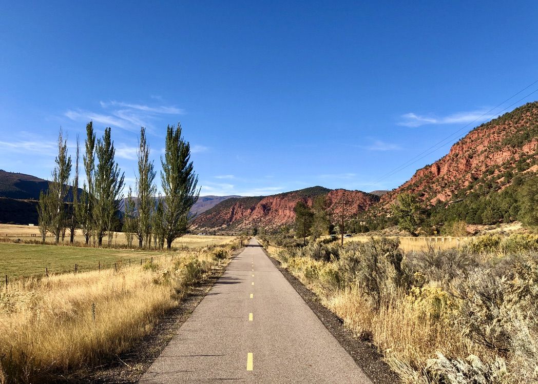 A paved path leads towards distant red mountains under a clear blue sky. - Carbondale – Satank Bridge loop from Carbondale