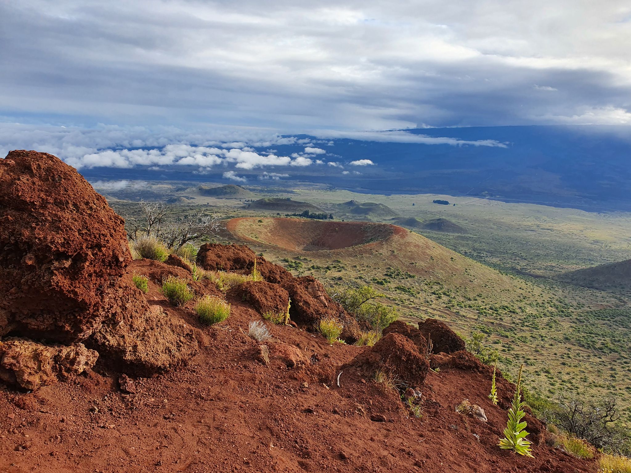 Red volcanic rocks in foreground, large crater, and cloudy distant landscape. - Pu‘ukalepeamoa Lookout