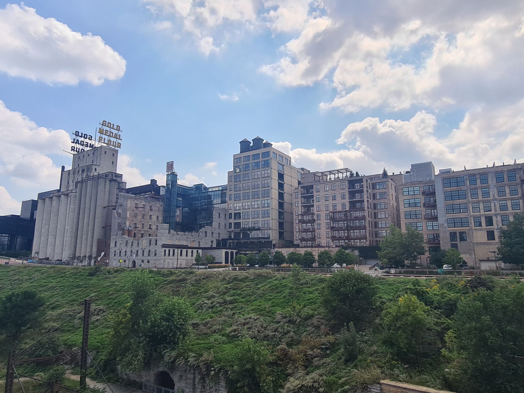 Historic industrial buildings and modern structures on a grassy hill under a cloudy sky. - Stone Arch Bridge – Boom Island Park loop from Fridley