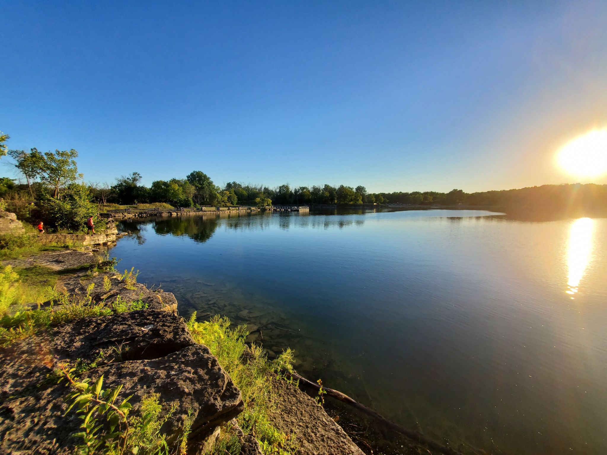Golden hour light on a calm lake with rocky banks, lush trees, and a person. - Menomonee Park Loop