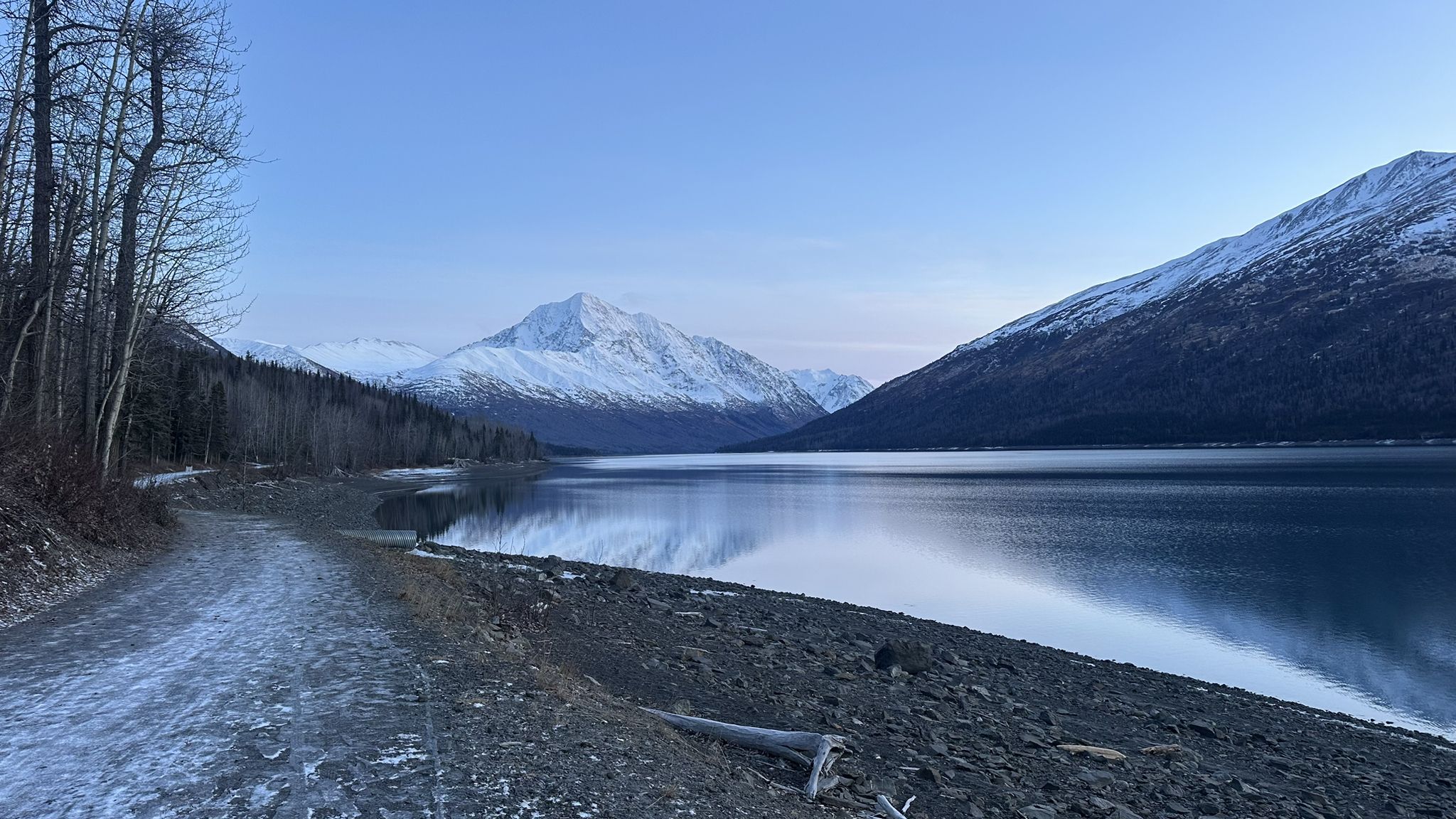 Calm lake reflecting snow-covered mountains under soft blue twilight, with a snowy path. - Eydlu Bena Loop Trail