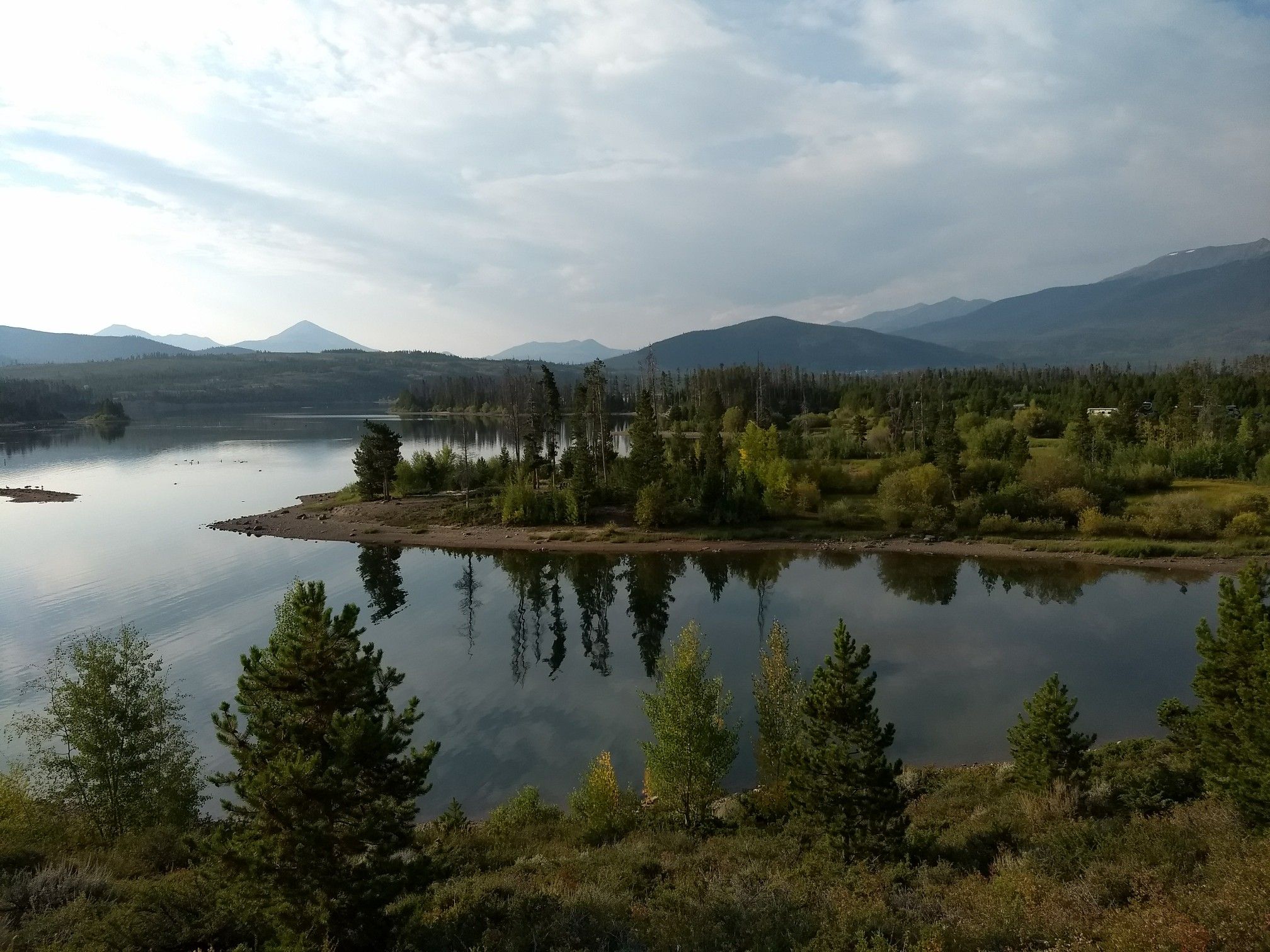 Calm lake reflecting trees and sky, with forested islands and mountains. - Old Dillon Reservoir Trail