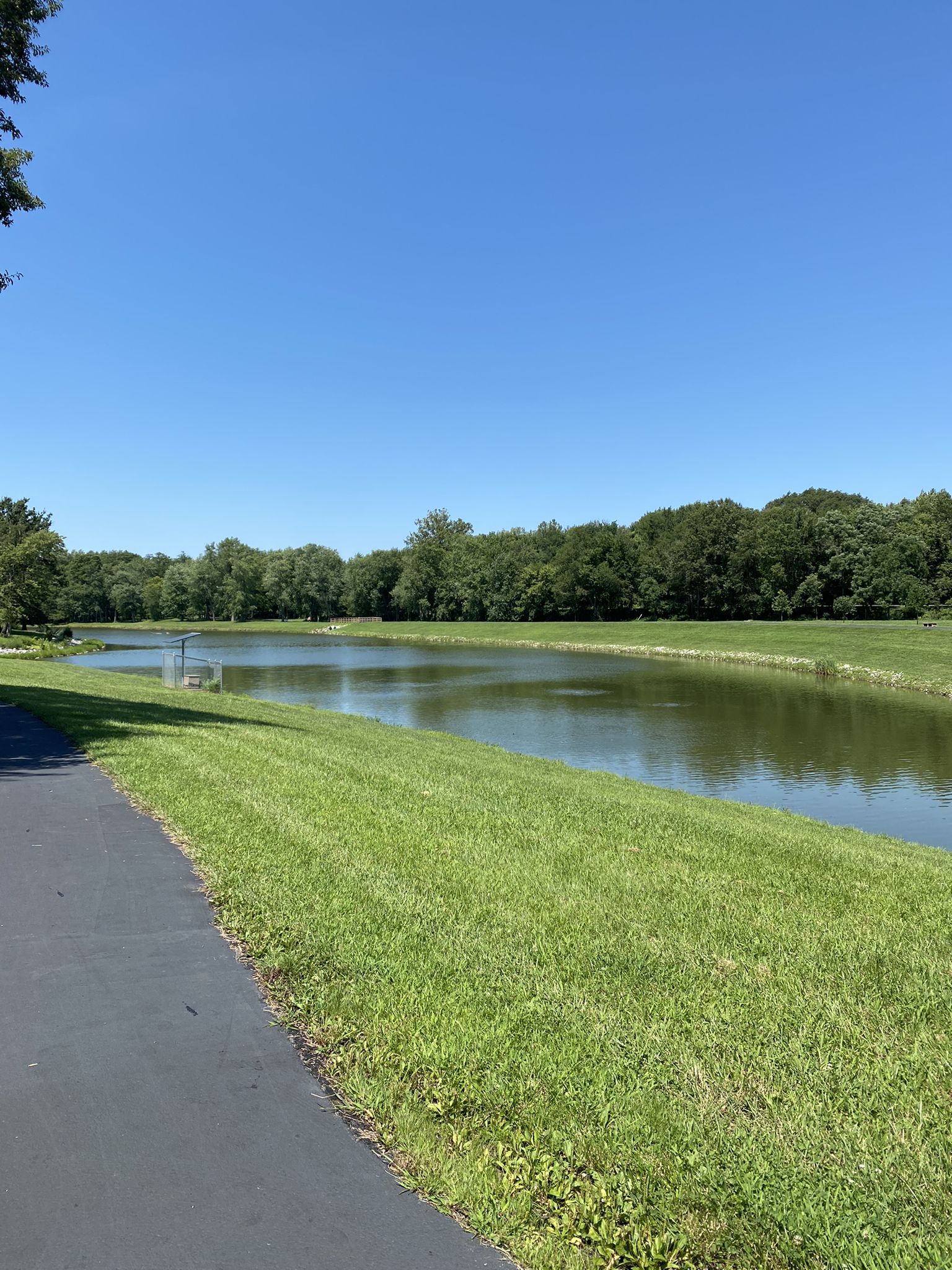 A calm pond with a grassy bank and paved path under a clear sky. - Heritage Trail
