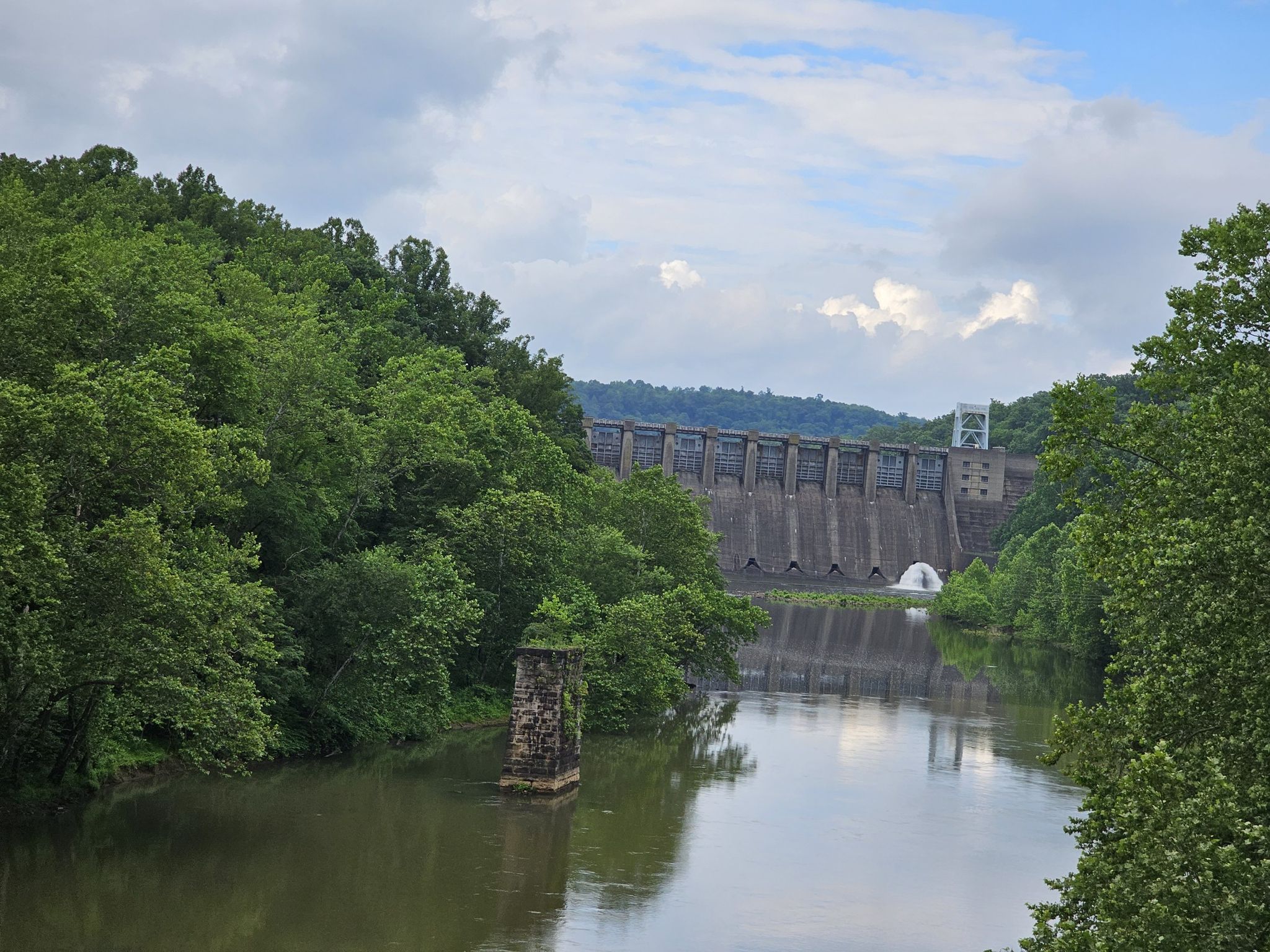 A river flows towards a large concrete dam, surrounded by dense green forest. - West Penn Trail