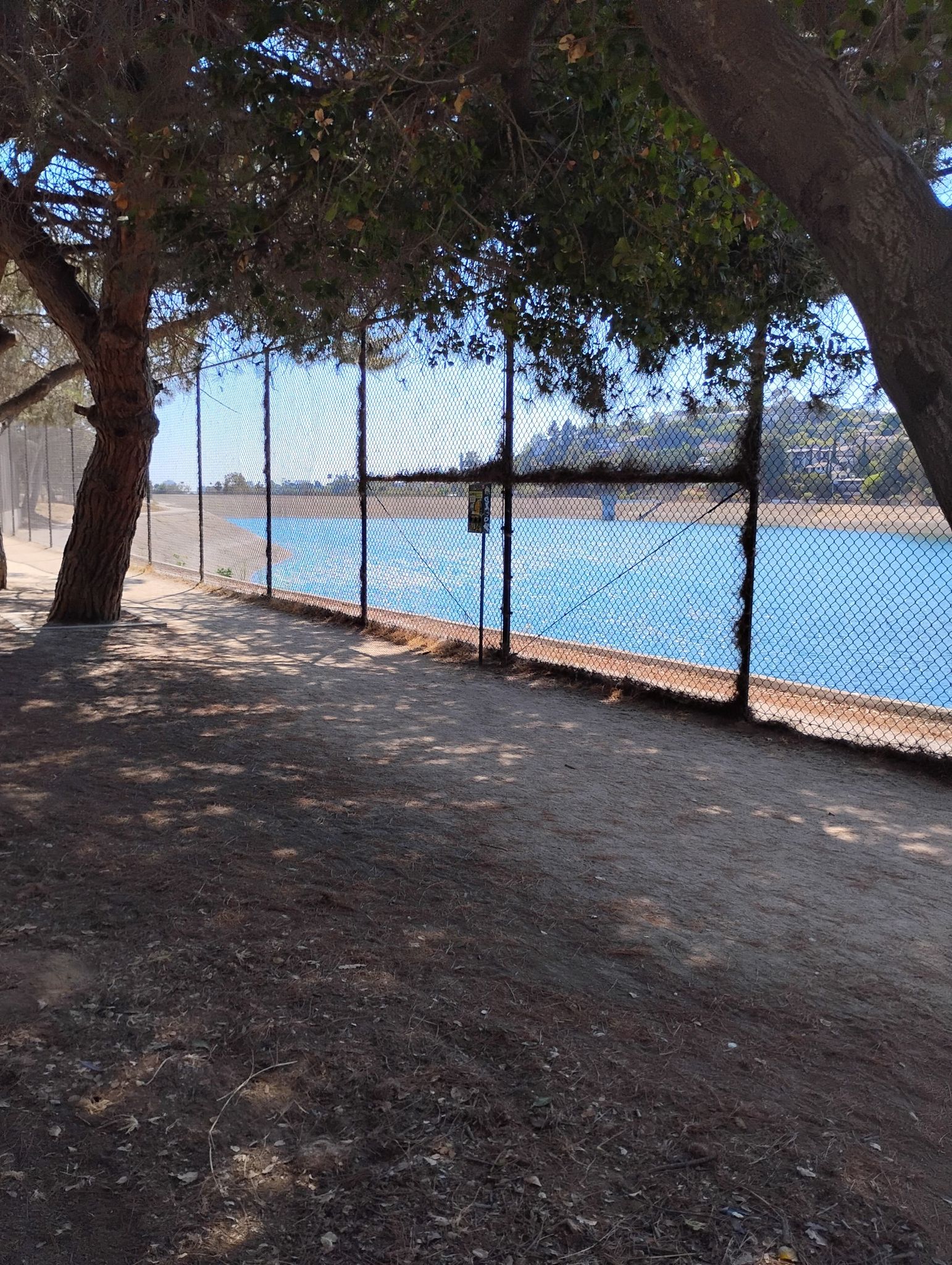 Sunny view of a dirt path, chain-link fence, and blue water with trees. - Silver Lake Reservoir Trail
