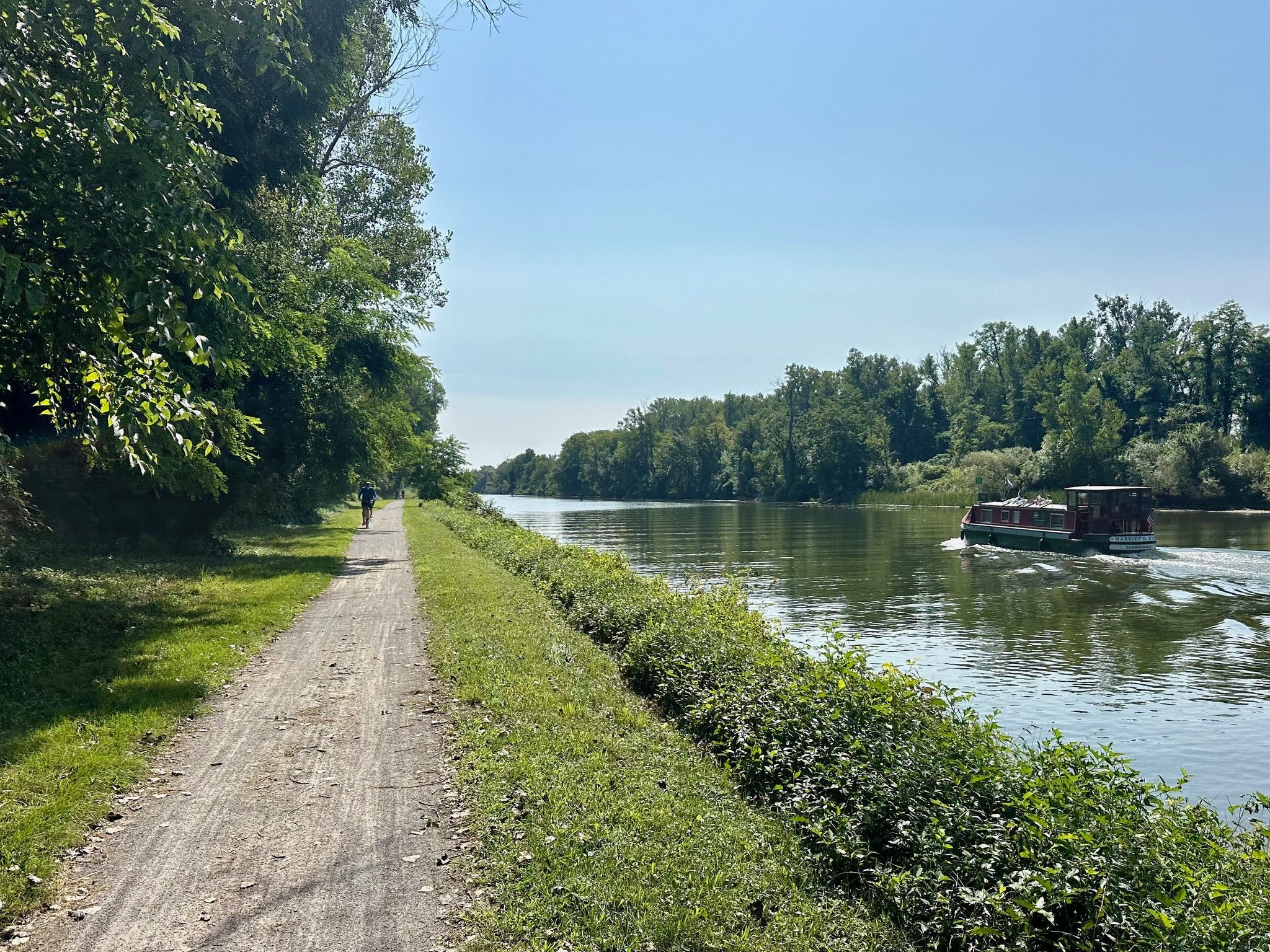 A person walks on a dirt path beside a river with a boat, sunny. - Erie Canalway Trail – Pittsford town loop from Oak Hill Country Club