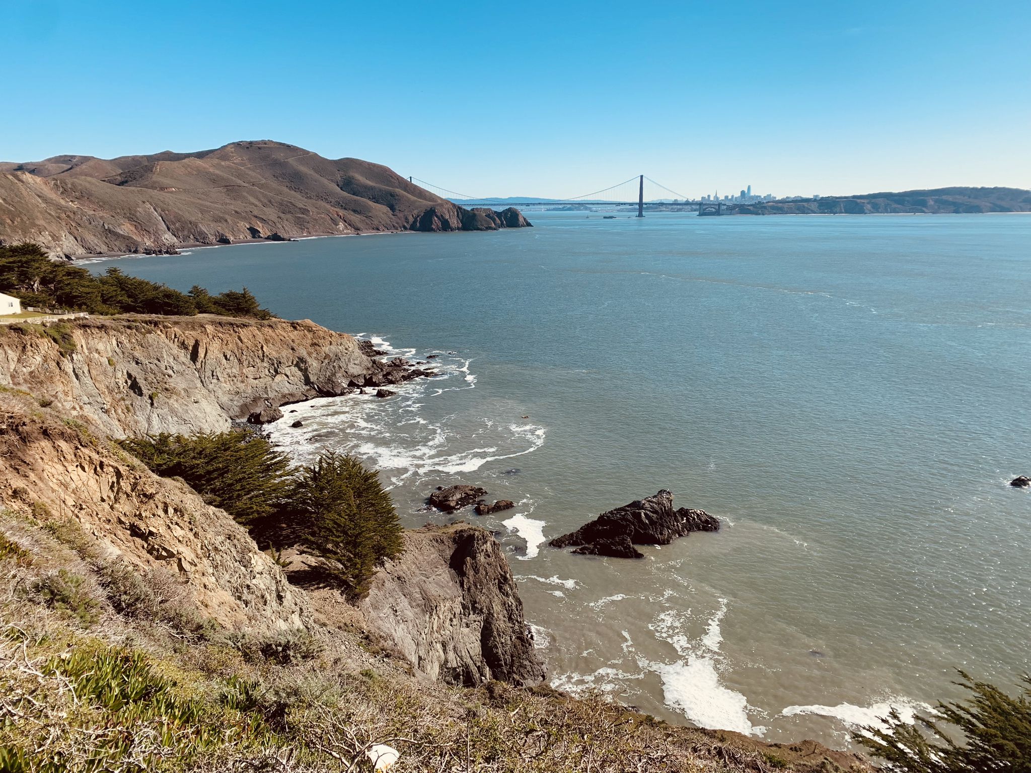 Rocky cliffs and coastline with waves, overlooking a wide bay and distant bridge. - Point Bonita Trail Loop