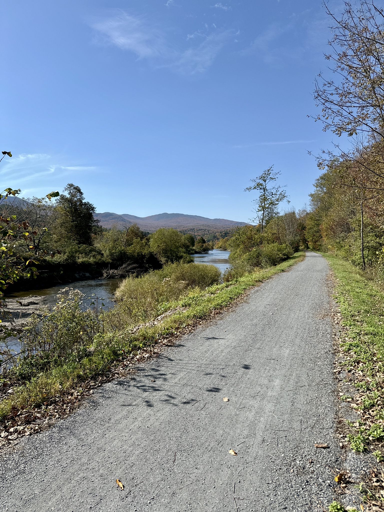 A gravel path runs beside a winding river with distant mountains and autumn trees. - Lamoille Valley Rail Trail