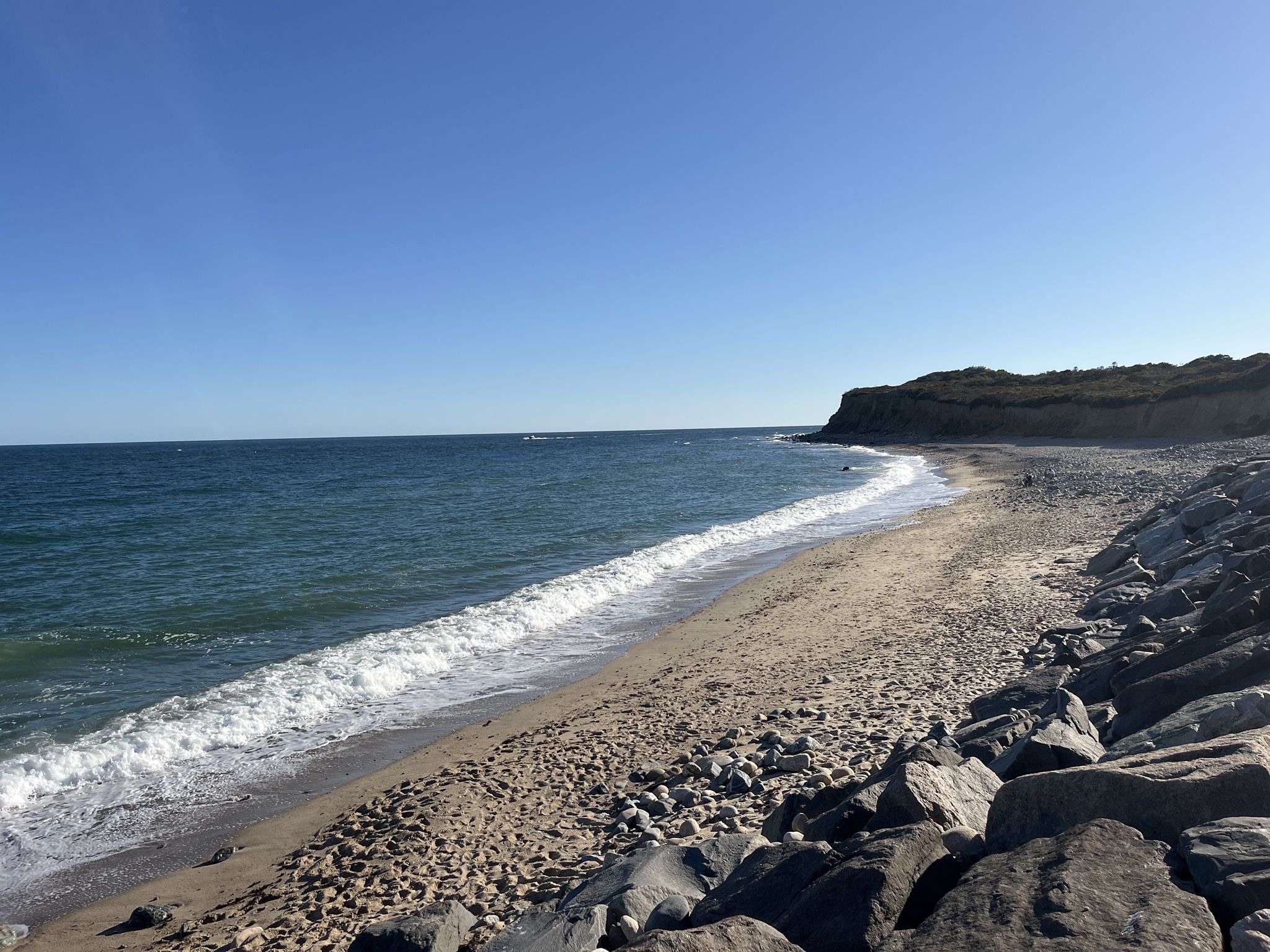 Sunny beach with waves breaking on sand and rocks, distant green cliffs. - Montauk Point State Parkway – Montauk Point Lighthouse loop from Montauk