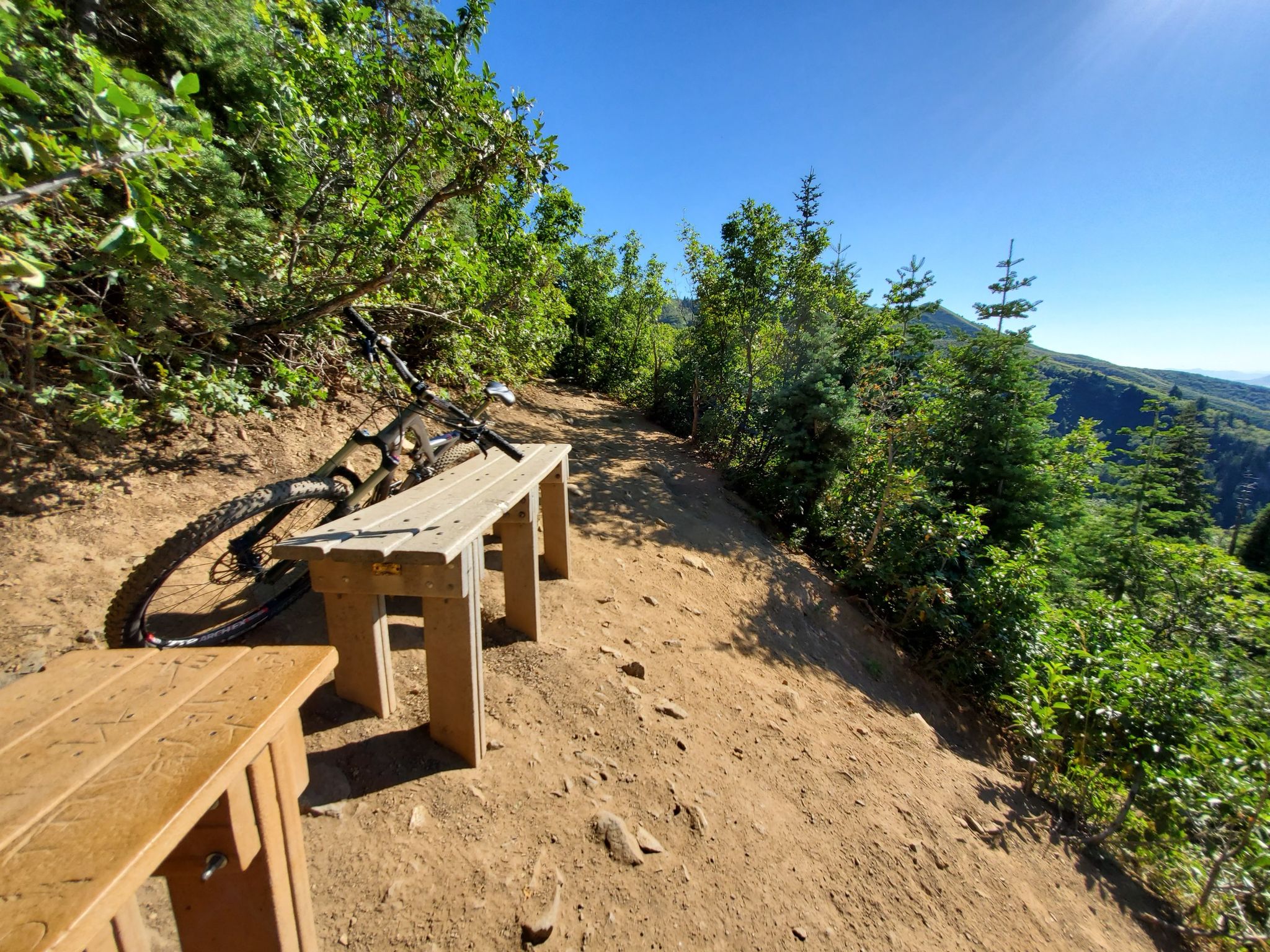 Mountain bike leaning on a wooden bench on a sunny dirt trail. - View of Mueller Park Trail – Big Rock loop from North Salt Lake