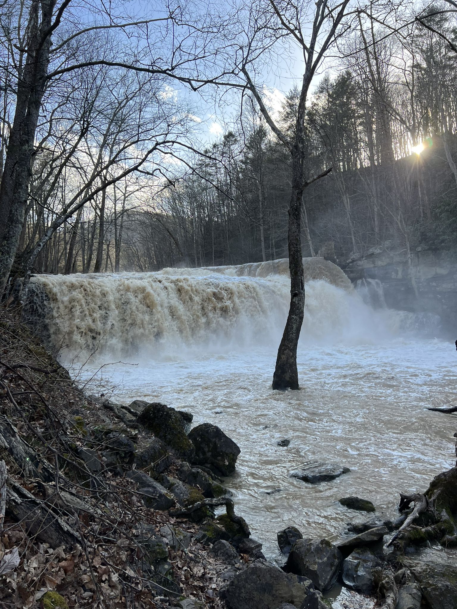 Muddy waterfall cascades over rocks in a forested landscape with bright sunlight. - Brush Creek Falls – White Oak Falls loop from Brush Creek Preserve