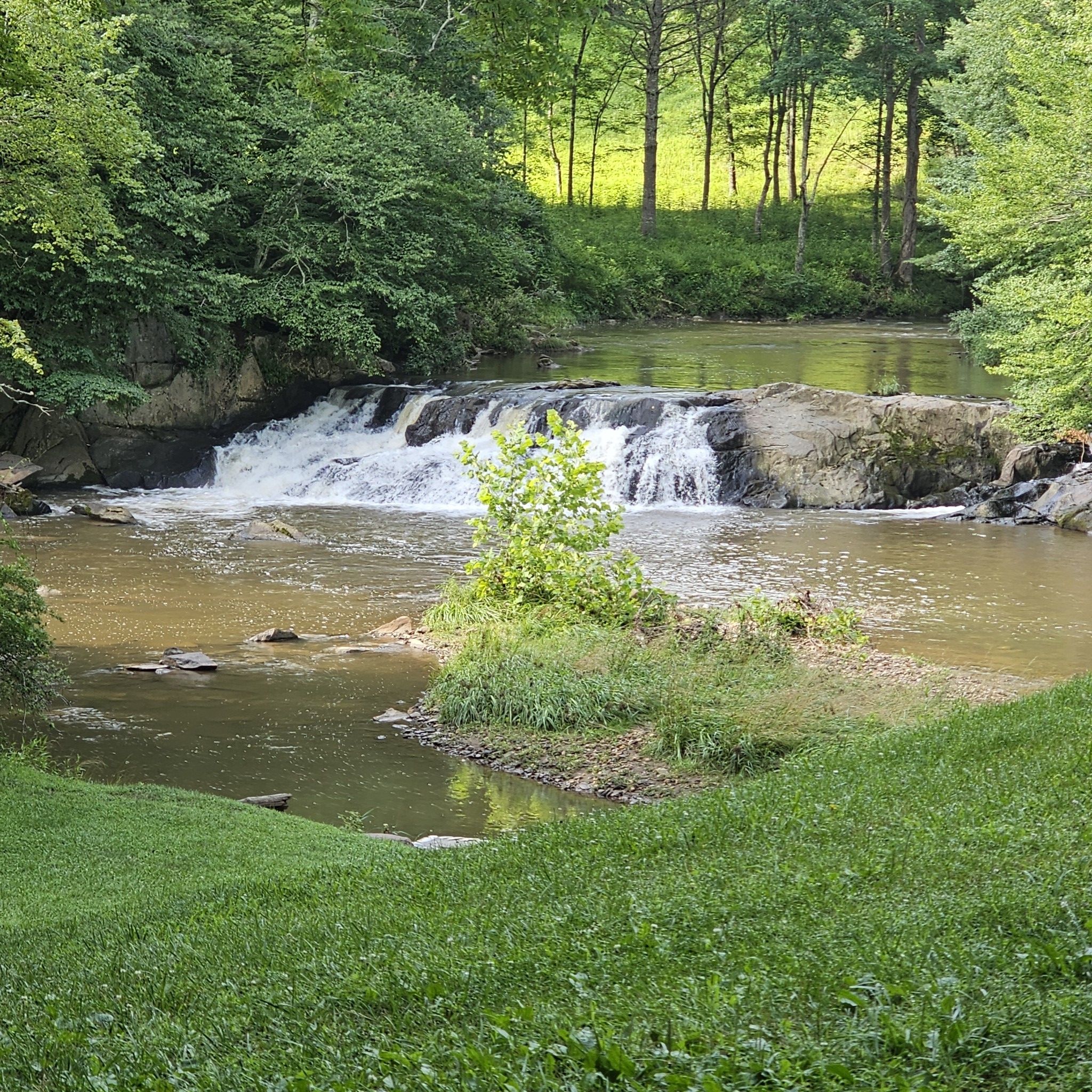Waterfall in a lush green forest with a wide river, bright daylight. - New River Trail: Galax to Fries Junction