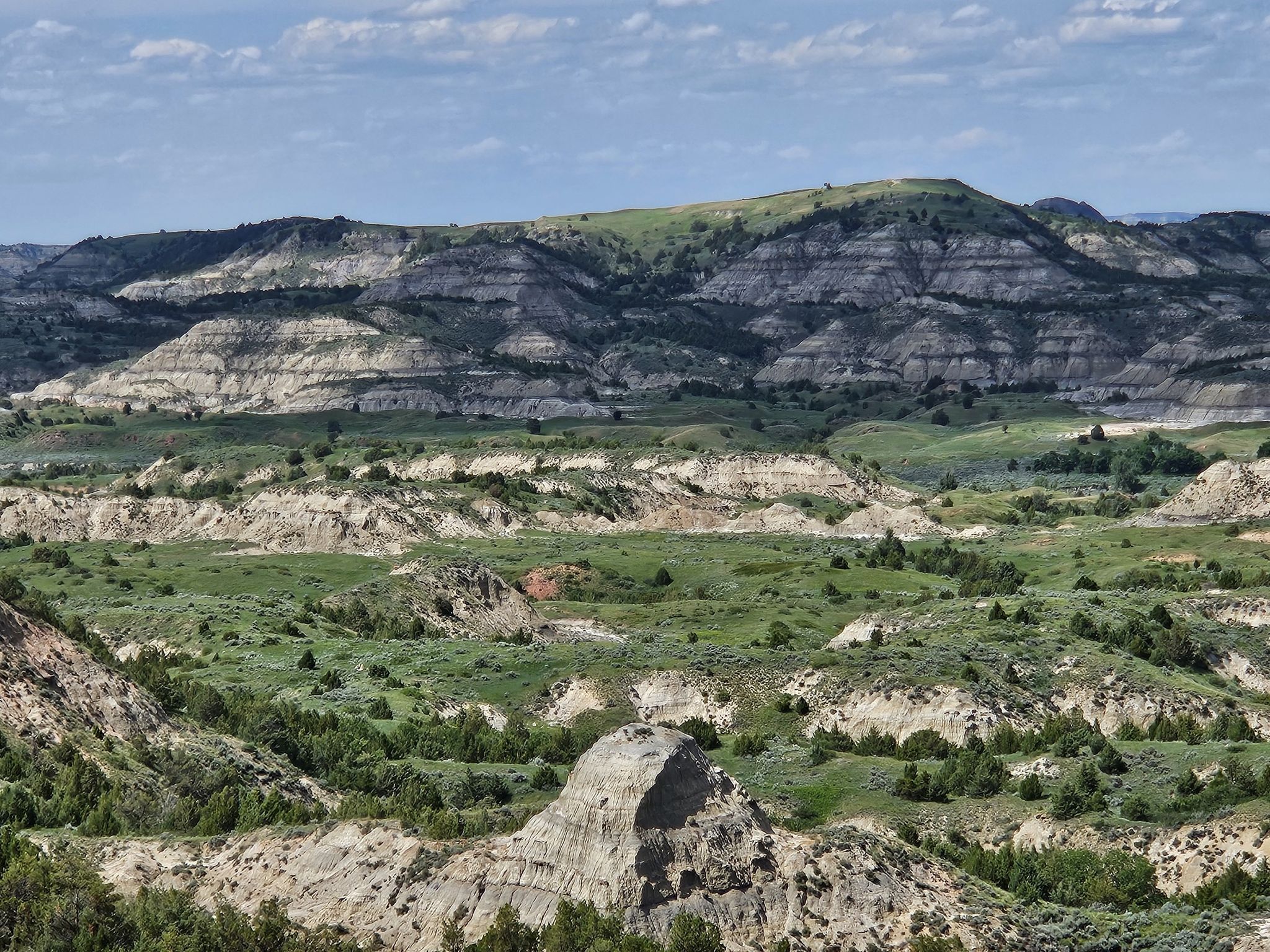 Layered badlands hills with green valleys under a partly cloudy sky. - Painted Canyon Trail