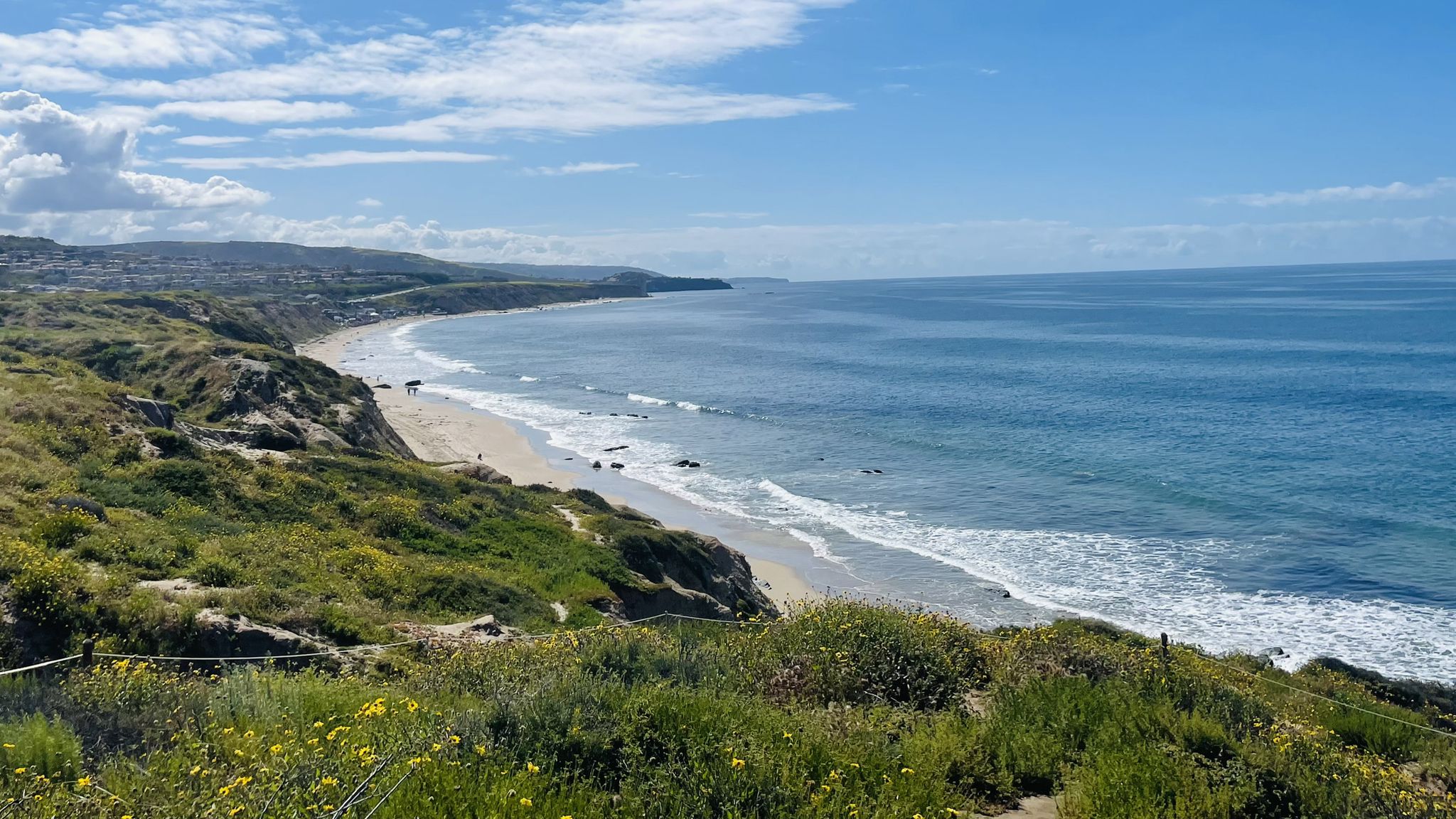 Sunny coastal view with green cliffs, sandy beach, blue ocean, and distant town. - Crystal Cove Trail