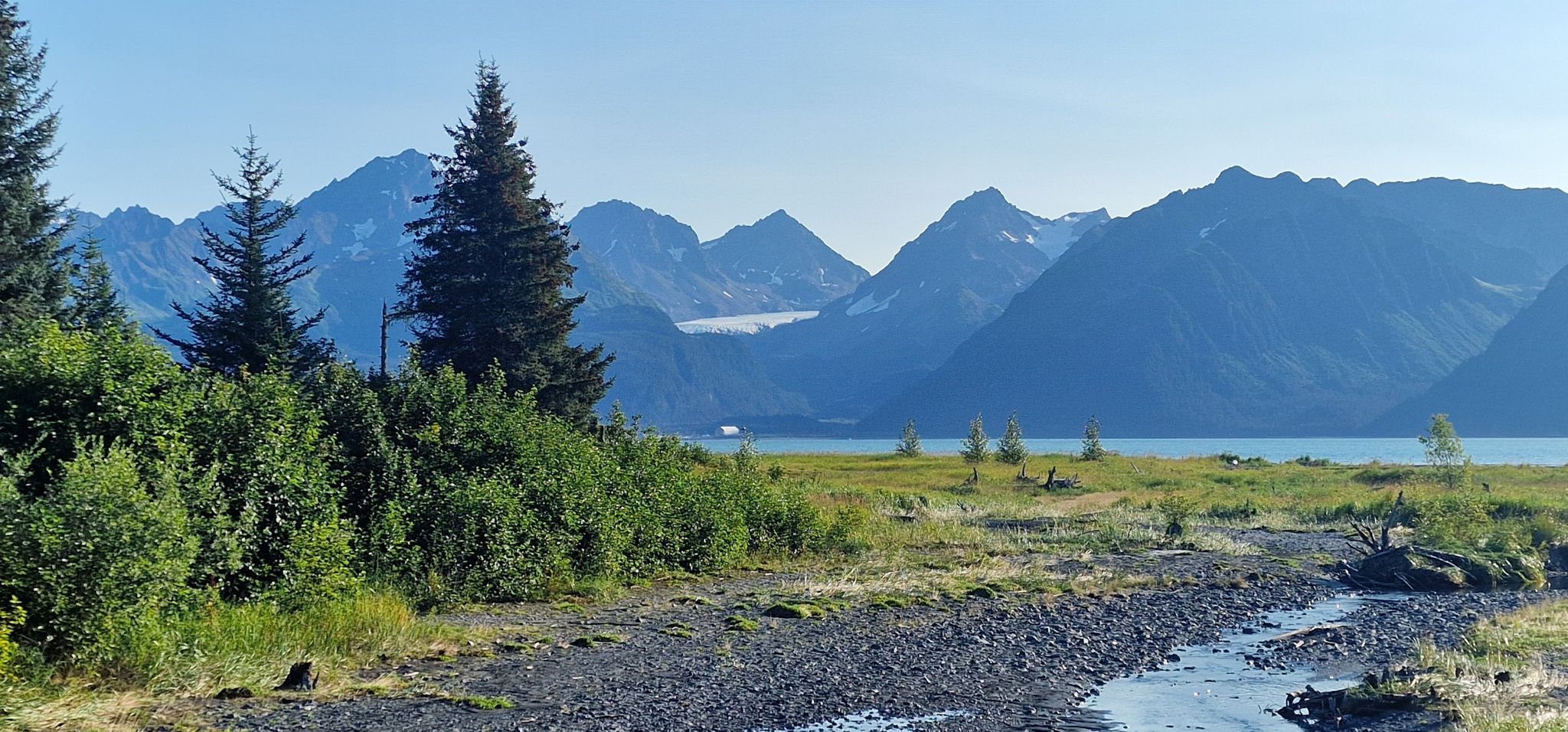 Stream flowing into a lake with forested hills and distant mountains under clear skies. - Caines Head Trail