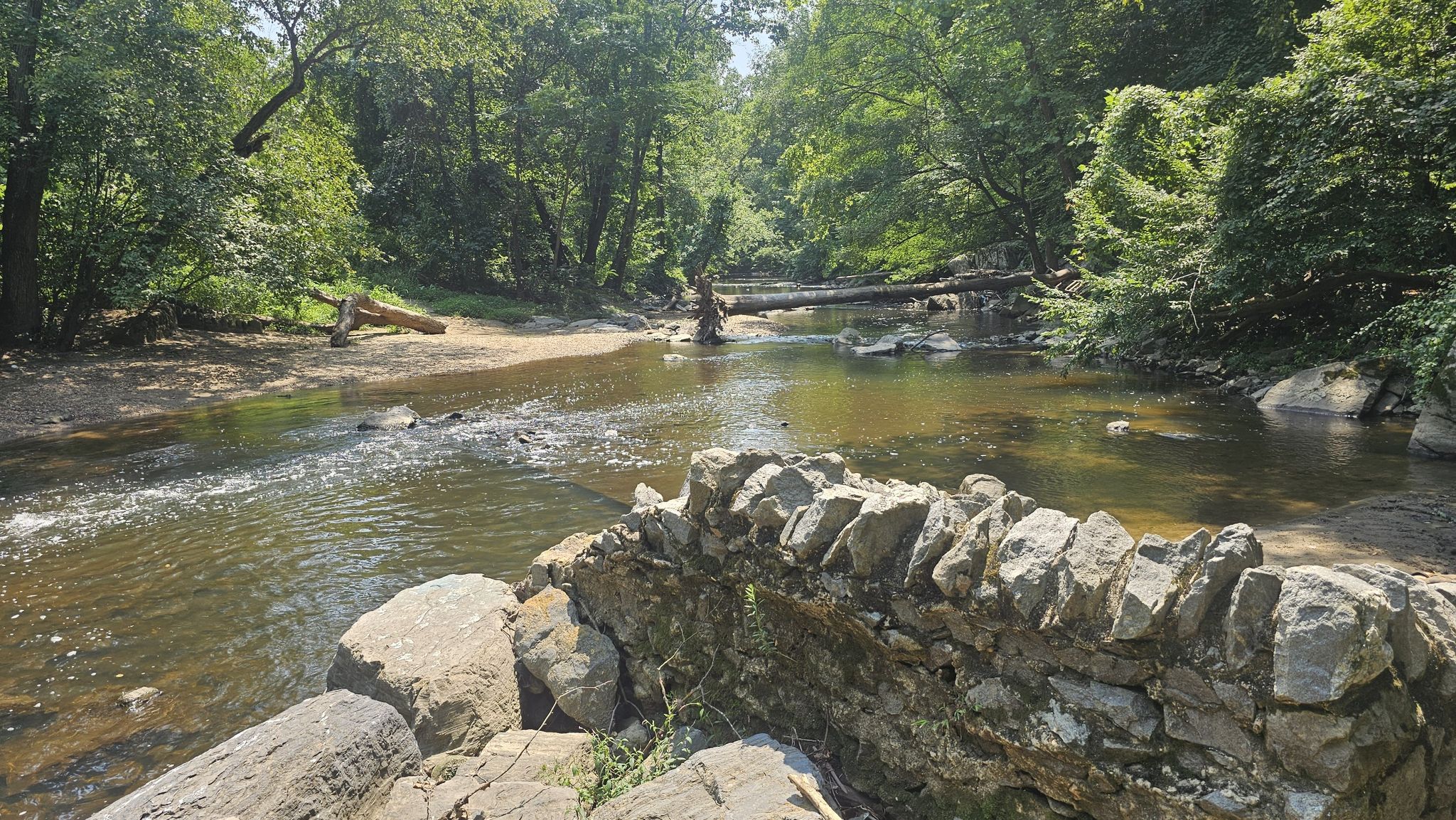 A rocky river with a stone wall in the foreground, surrounded by green trees. - Milkhouse Ford Loop