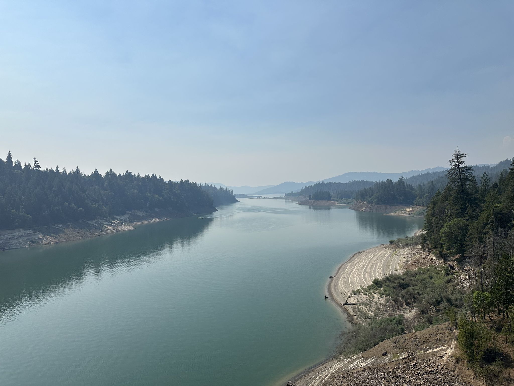 A wide, calm river flows between forested banks under a hazy sky. - Rogue Gorge via Rogue-Umpqua Scenic Byway