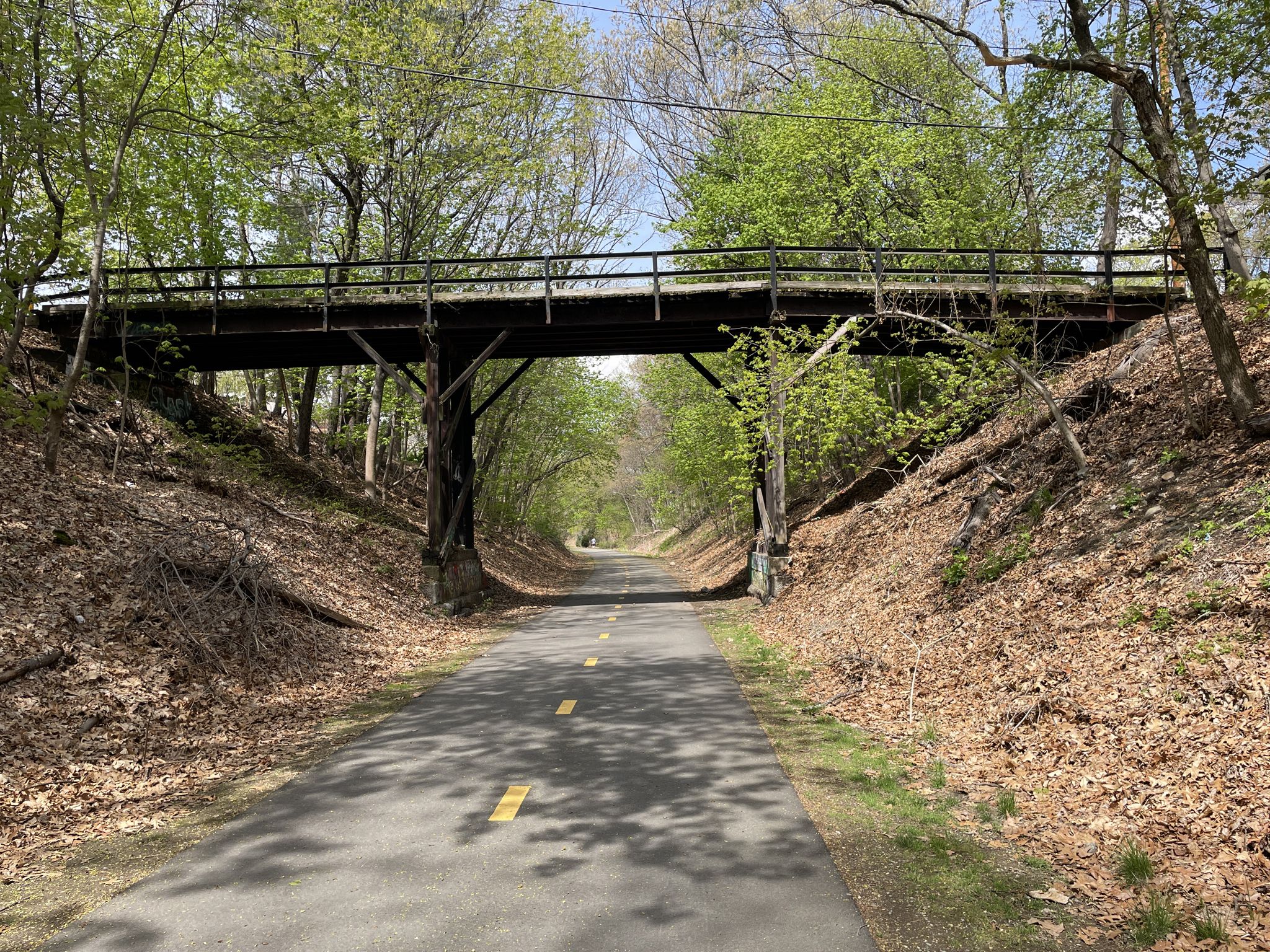 Paved path under a rustic wooden bridge, surrounded by green trees. - Cochituate Rail Trail – Lake Cochituate (Middle Pond) loop from Jordan's Furniture