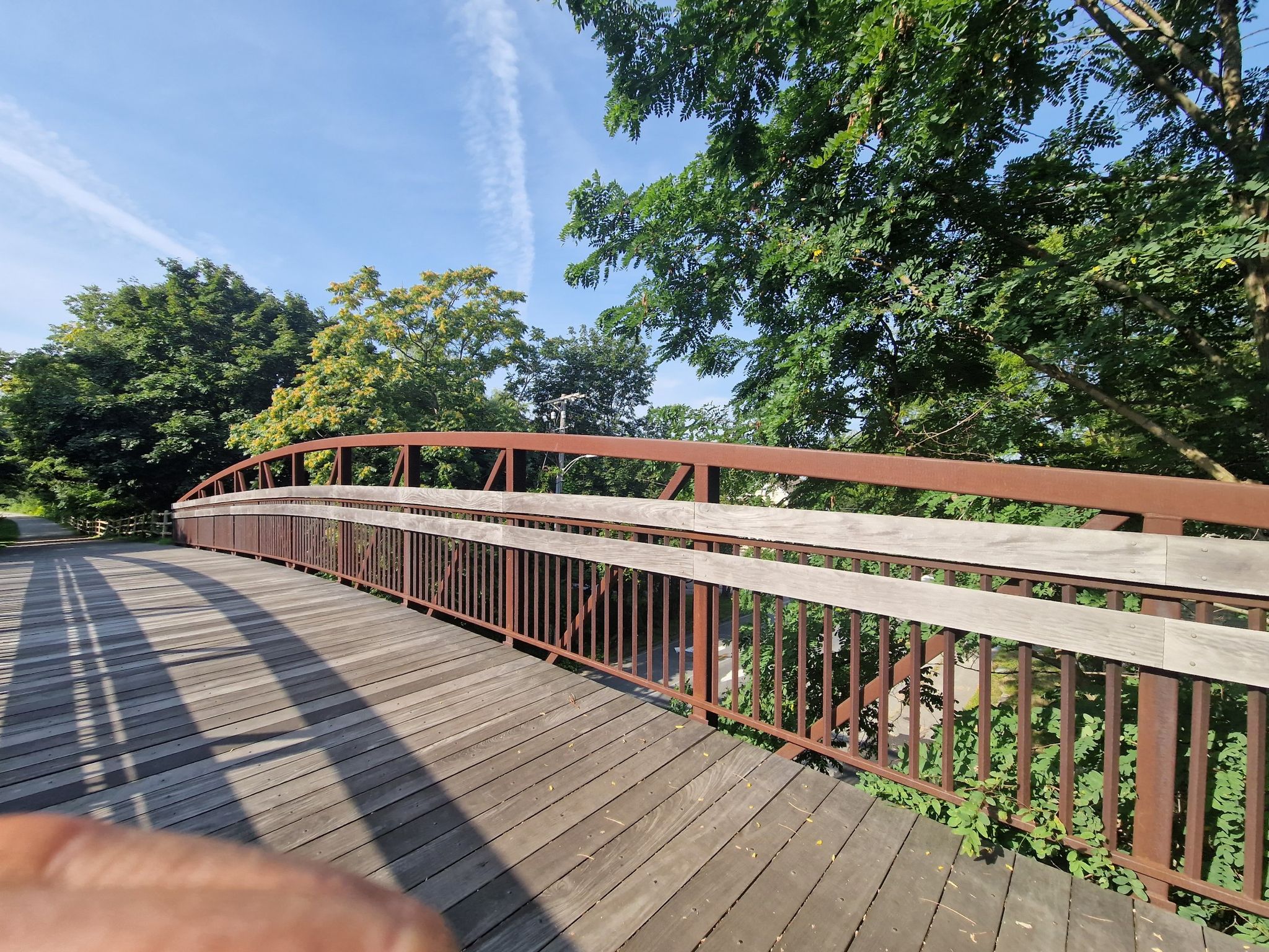Wooden and metal pedestrian bridge surrounded by green trees on a sunny day. - Clipper City Rail Trail
