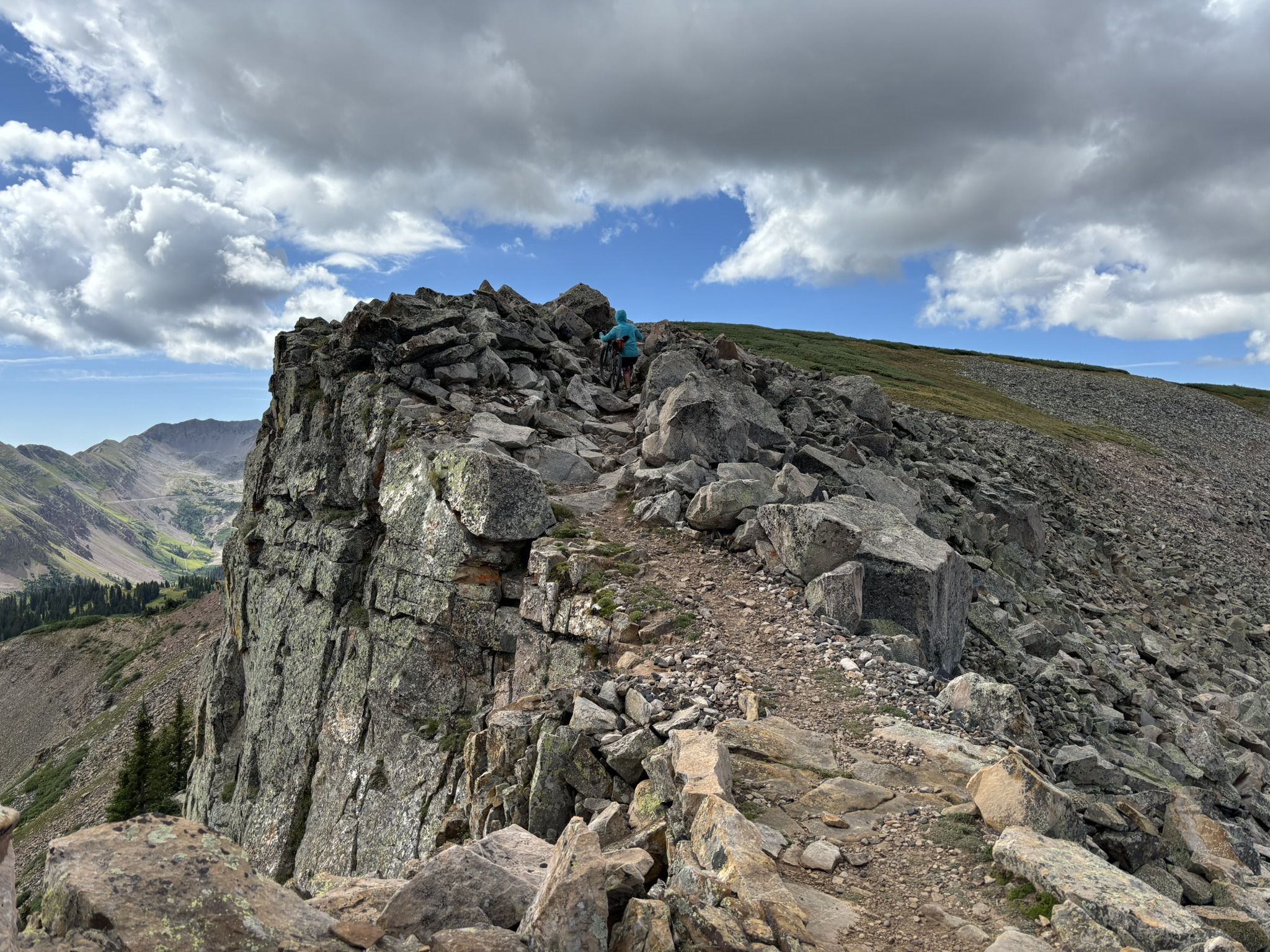 A person hikes a narrow, rocky mountain ridge under a cloudy sky. - San Juan Skyway