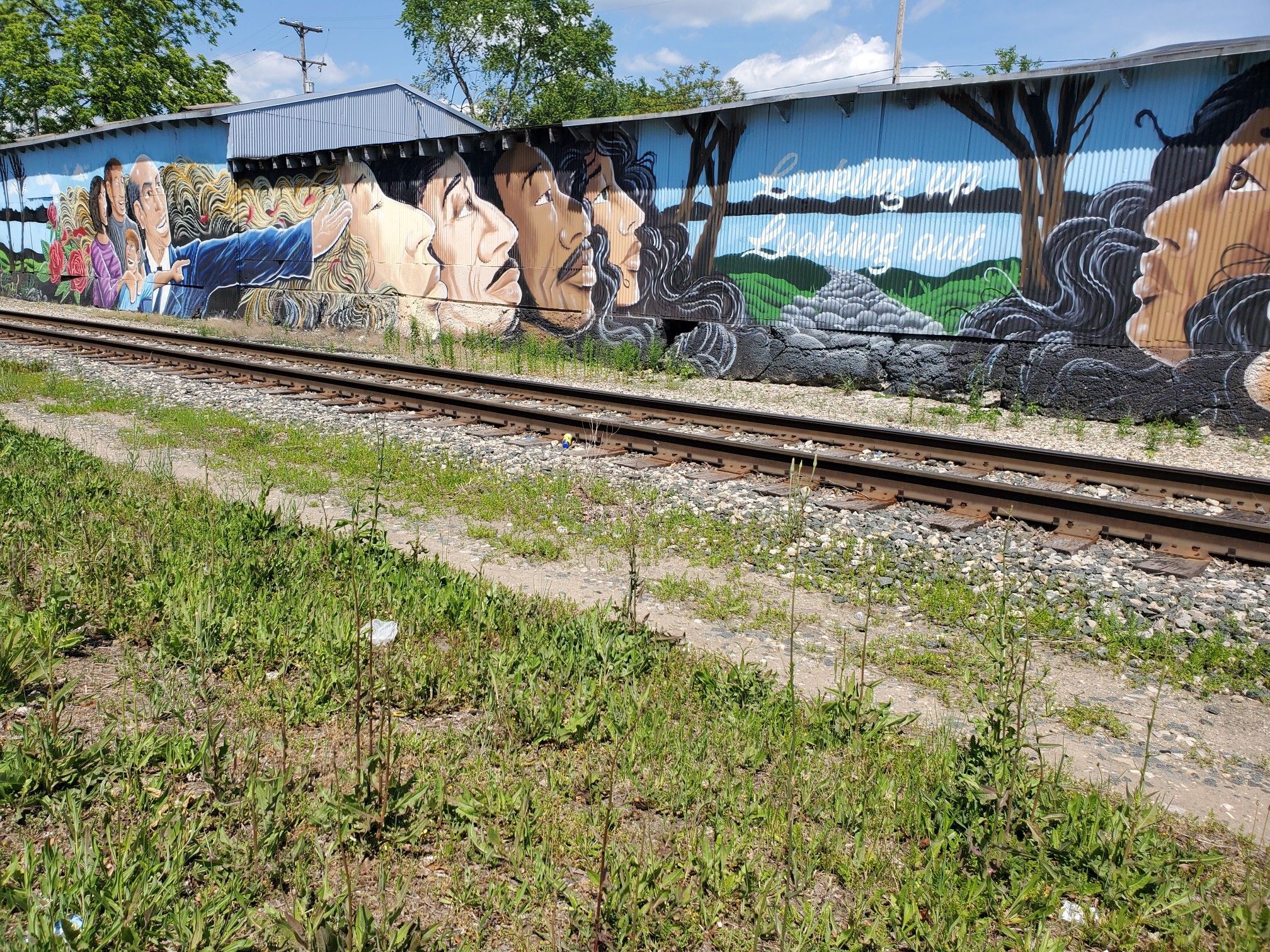Bright mural of faces and text on a wall beside railroad tracks in daylight. - Fred Meijer Standale Trail and Millennium Park Loop