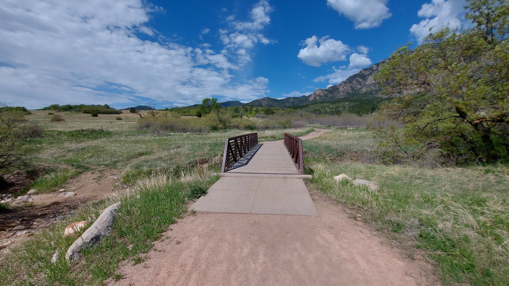 A sunny view of a metal bridge crossing a grassy field towards mountains. - Cheyenne Mountain State Park loop from Cheyenne Mountain State Park