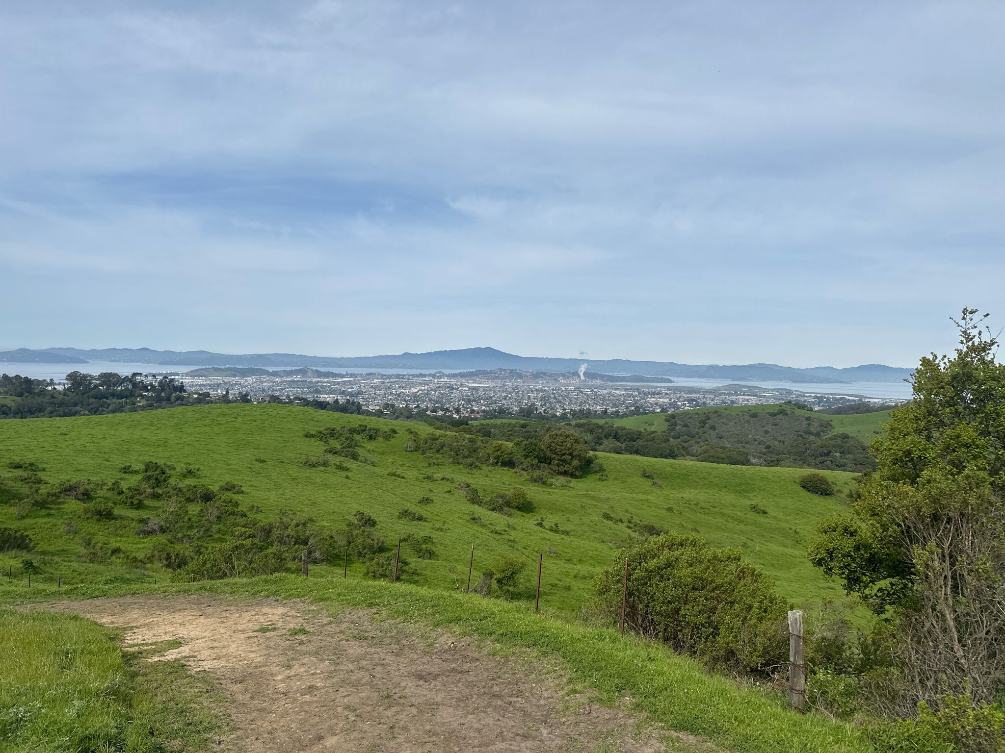Dirt path overlooking lush green hills and a distant city under a cloudy sky. - Leonards and Mezue Trail Loop