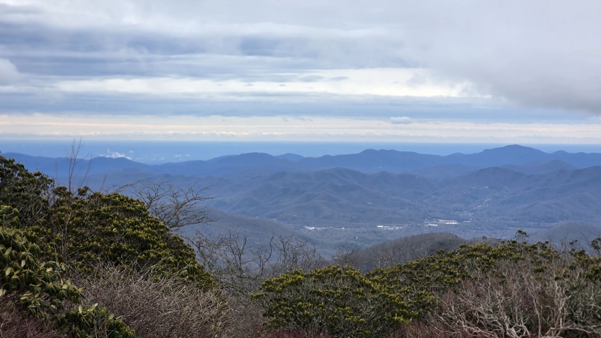 Overcast view of layered blue mountains and distant horizon, with green bushes. - Craggy Gardens Picnic Area – Craggy Gardens viewpoint loop from Bearpen Knob