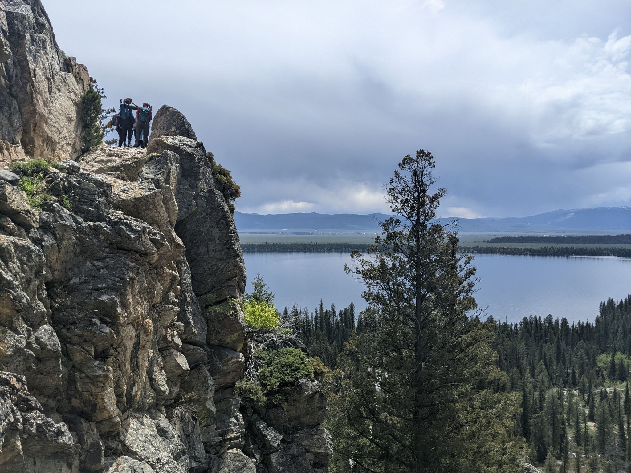 People on a rocky cliff overlooking a large lake and forest under cloudy sky. - View of Jenny Lake – Cascade Creek loop from Jenny Lake