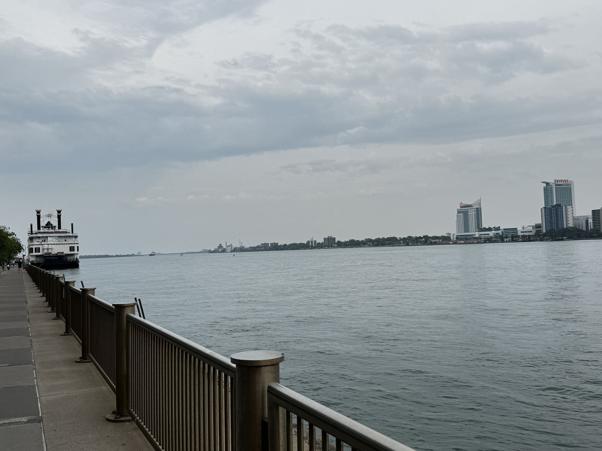 Waterfront promenade with a fence, river, and city buildings in the distance. - Detroit Riverwalk and Corktown Loop