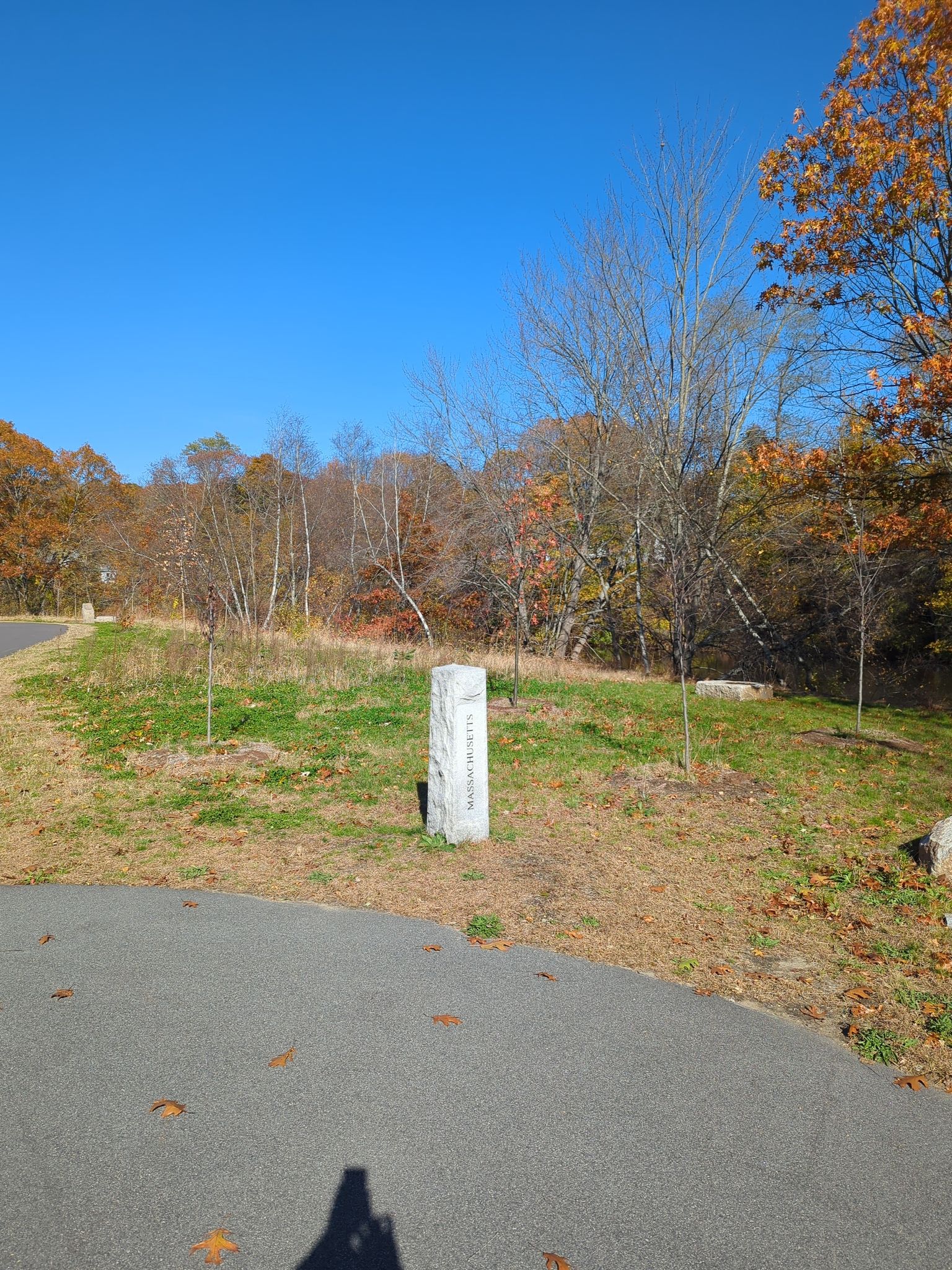 A white stone marker stands by a paved path with autumn trees. - Blackstone River Bikeway