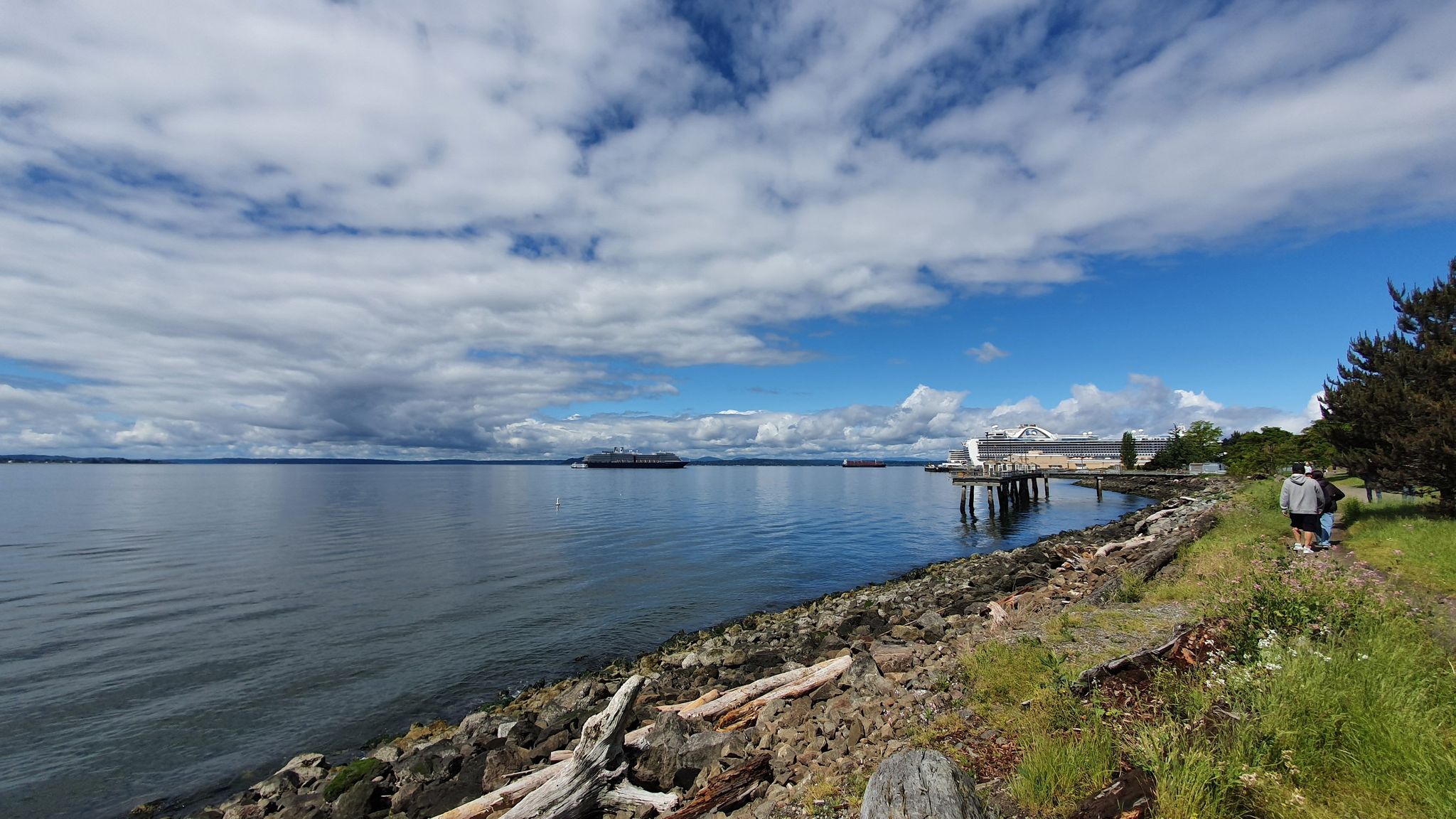 Calm bay with distant ship, rocky shore, and two people walking under cloudy sky. - Elliott Bay Trail