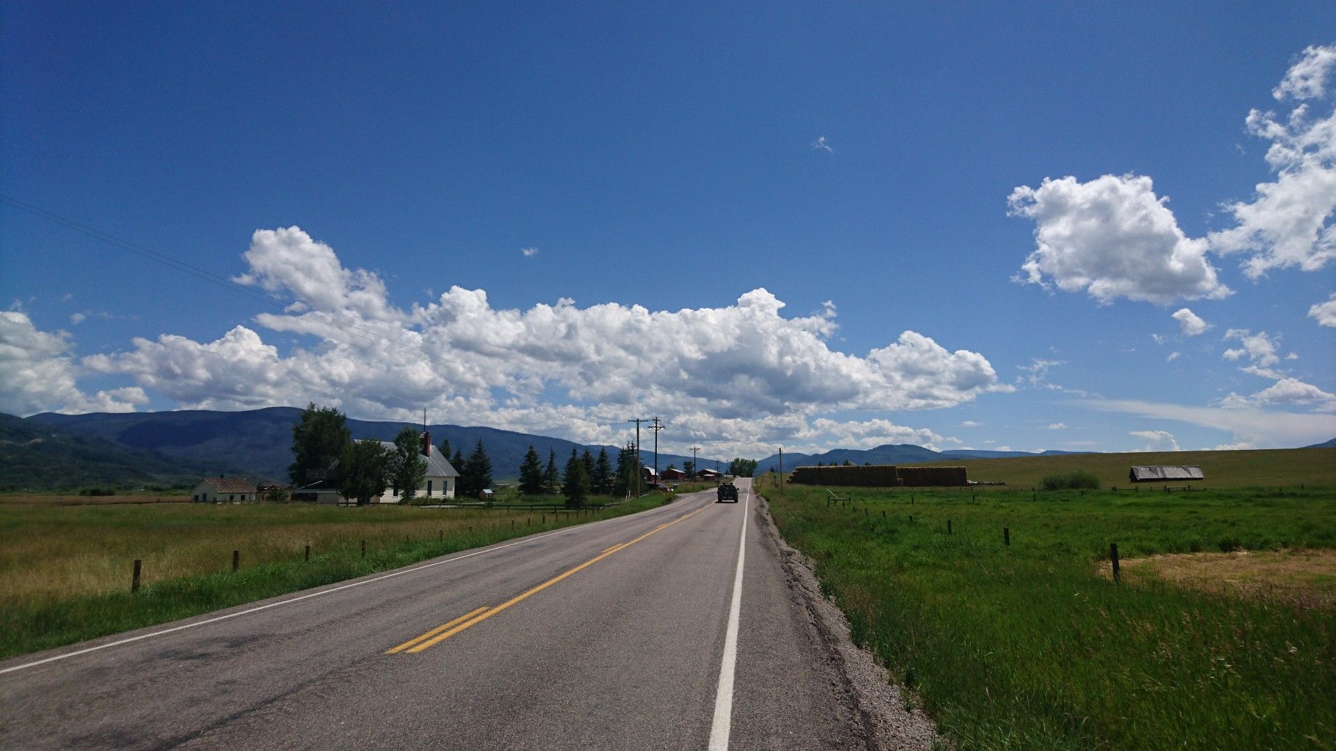 A paved road stretches through green fields with distant buildings under a cloudy blue sky. - Stagecoach/Oak Creek loop