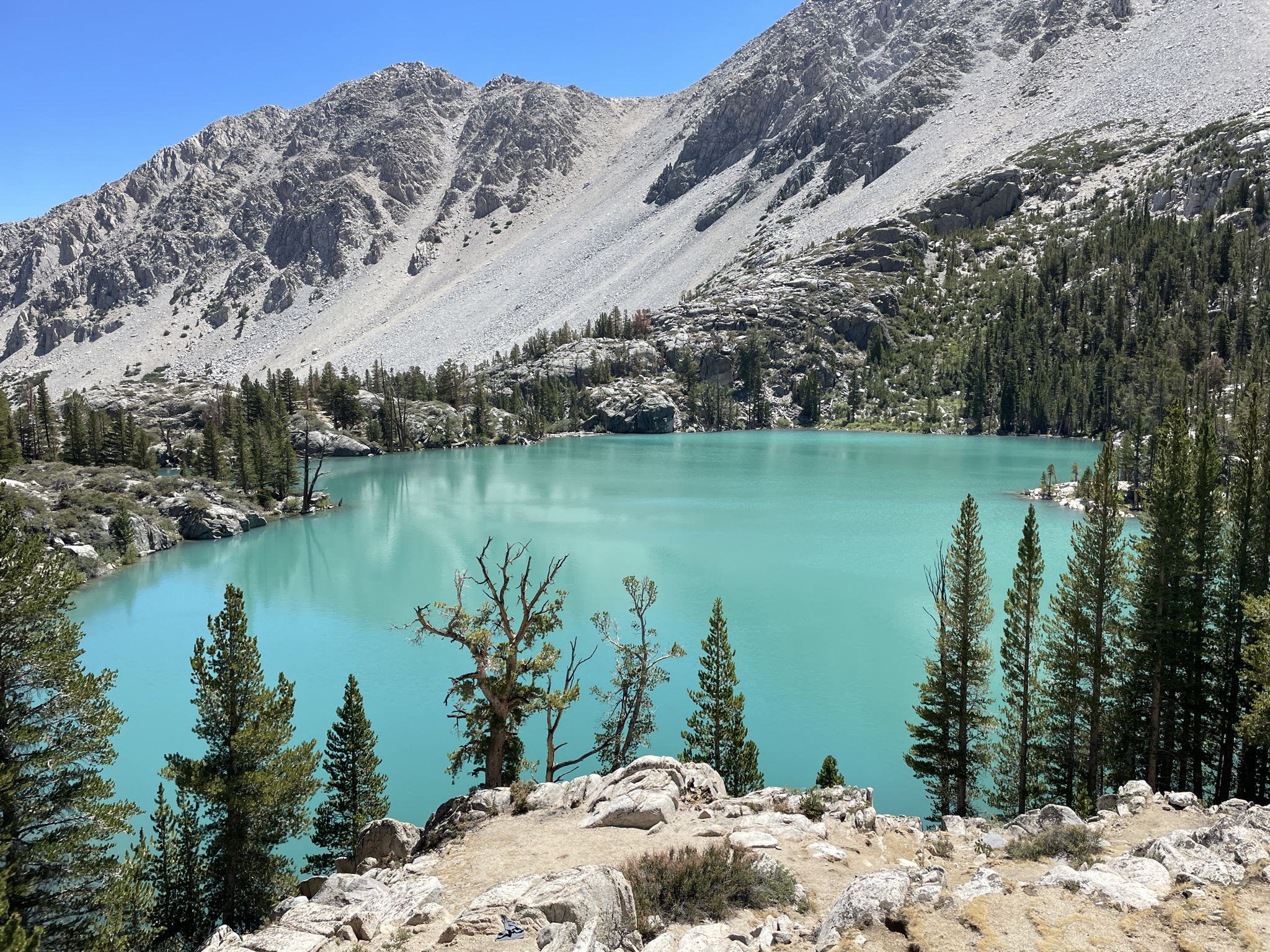 Bright turquoise mountain lake with rocky slopes and pine trees under clear sky. - Big Pine Lakes Trail