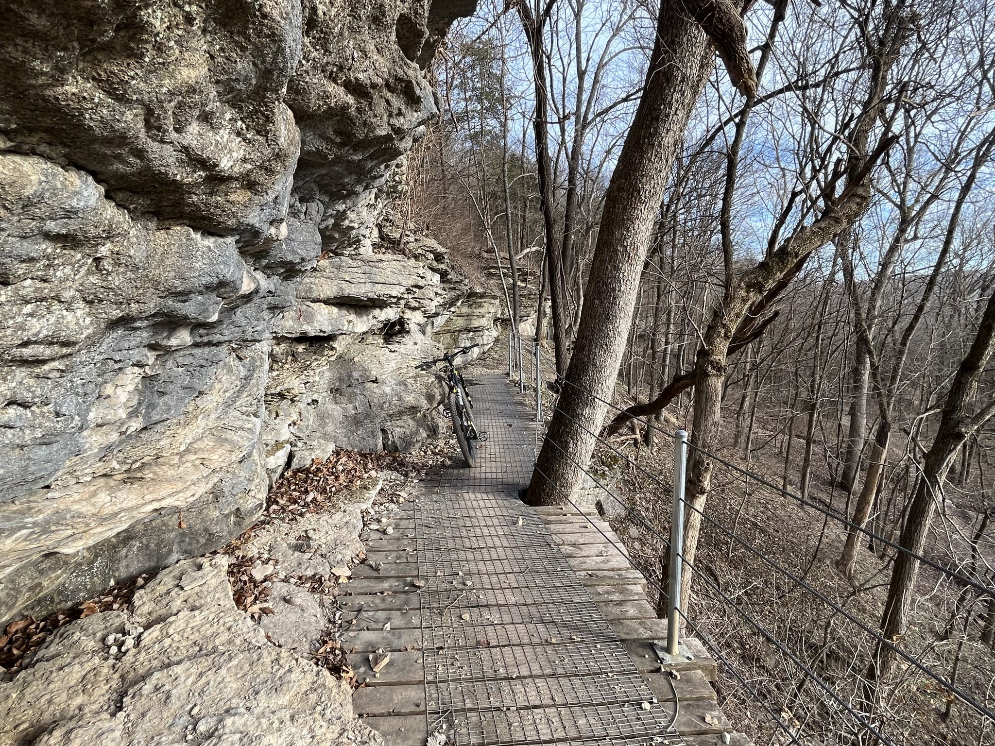 A wooden boardwalk with railings built into a rocky cliffside in a bare forest. - Wonderland Trails RV Hub – Bentonville City Square loop from Bentonville
