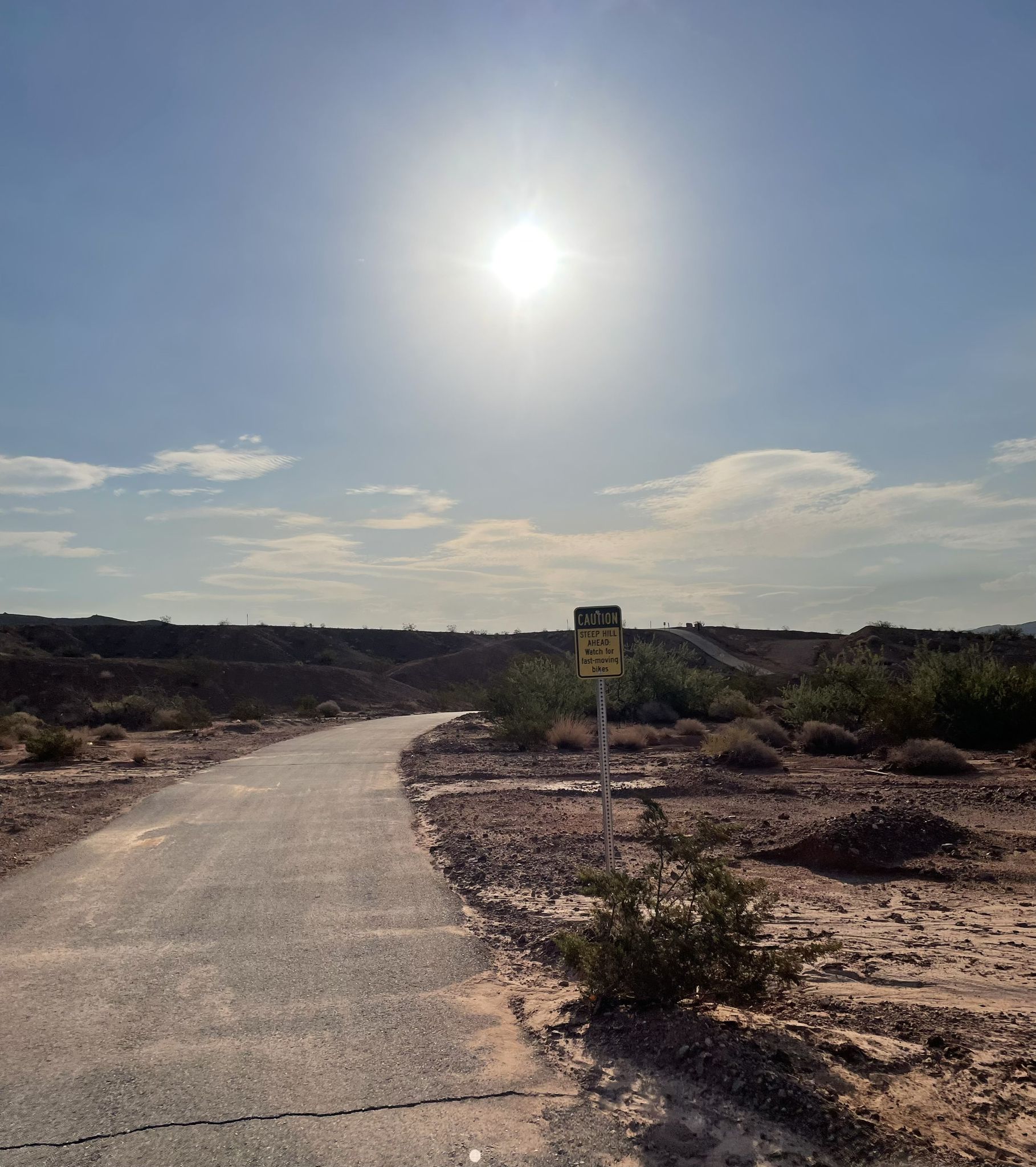 A paved path winds through a sunny, arid desert landscape with distant hills. - Wetlands Trail Short Loop
