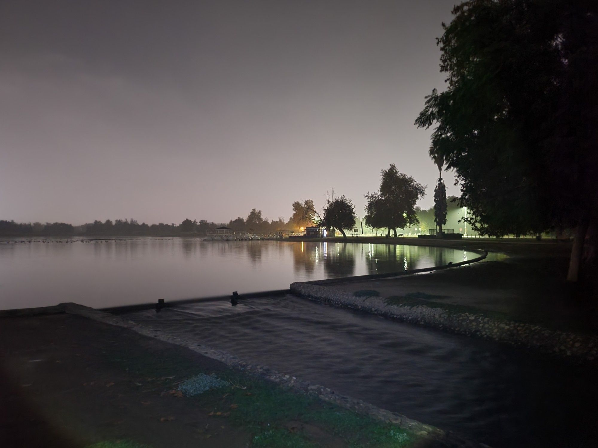 Dark lake at night with trees and distant lights reflecting on the water. - Lake Balboa Trail