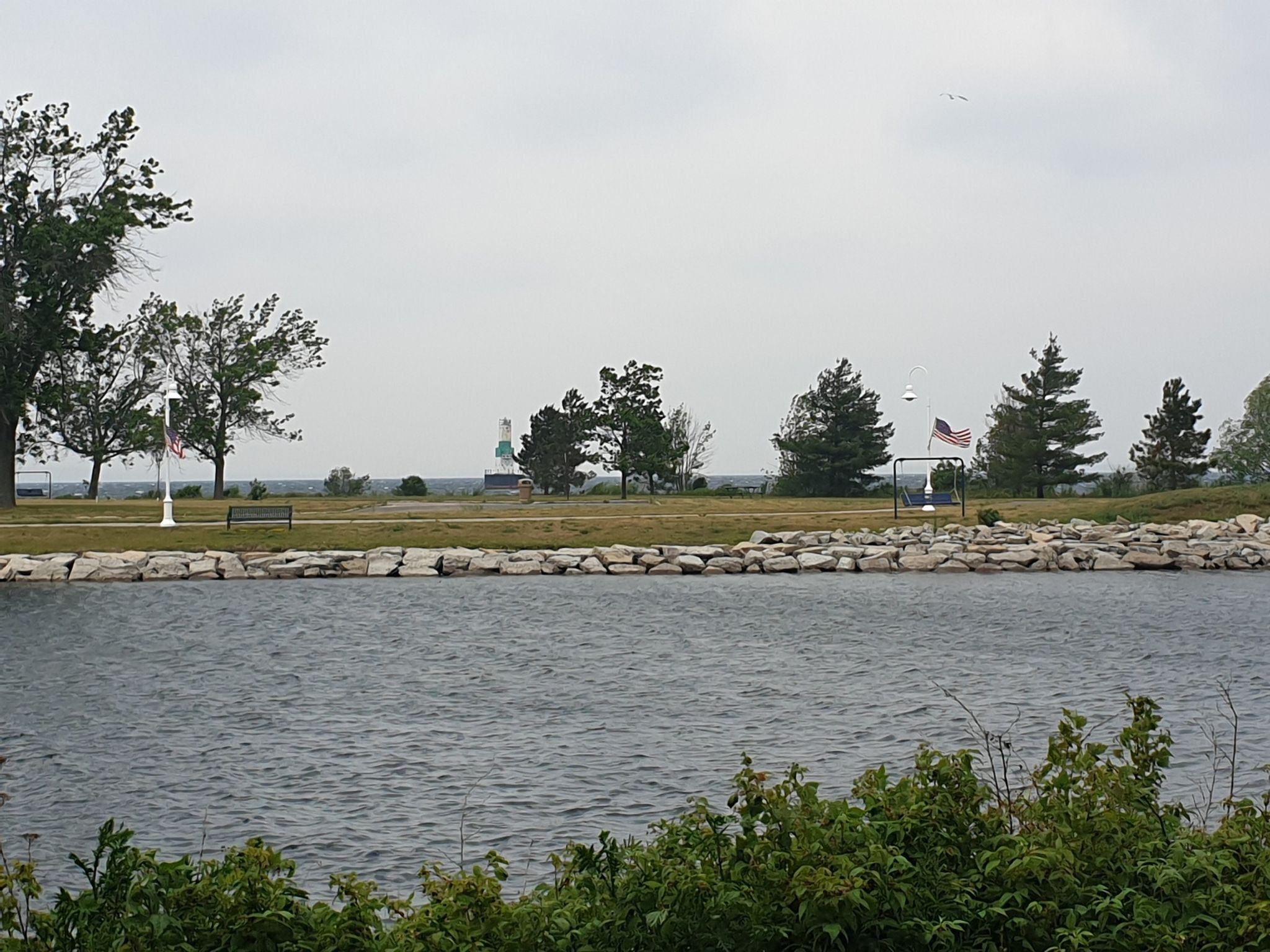 Calm water, rocky shore, and a park with trees under an overcast sky. - Ludington Park and Aronson Island Loop
