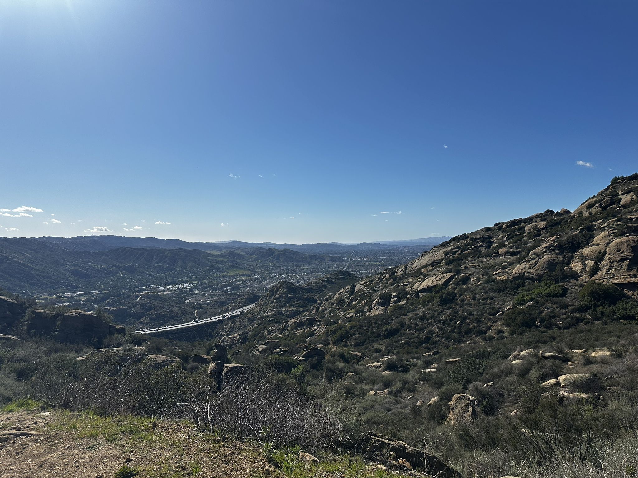 Sunny view of a rocky, brush-covered hillside overlooking a distant valley and city. - Wildlife Corridor Trail