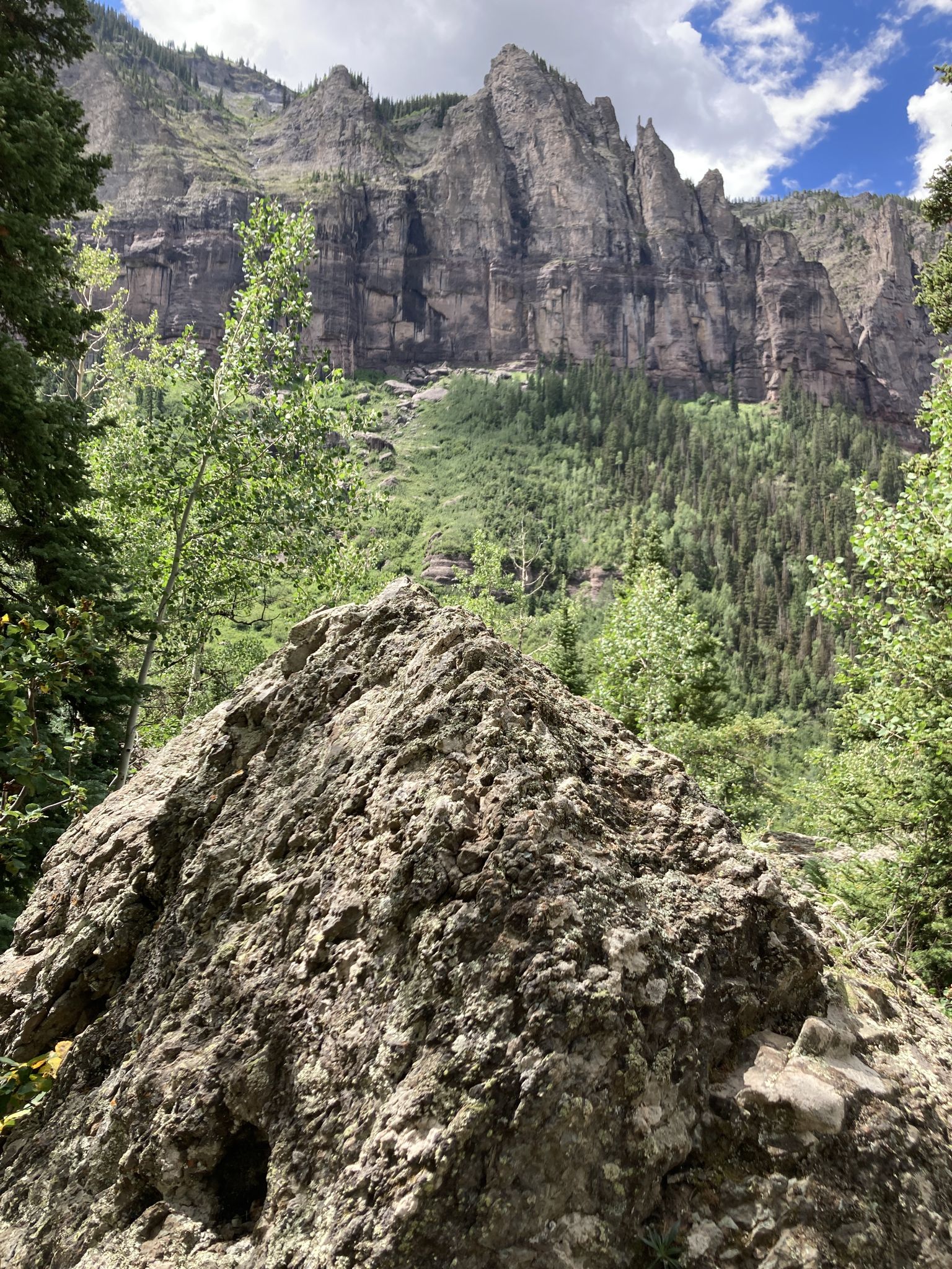 Large rock in foreground with towering, rugged cliffs and green trees behind. - Bridal Veil Falls to Ajax and Telluride Peaks