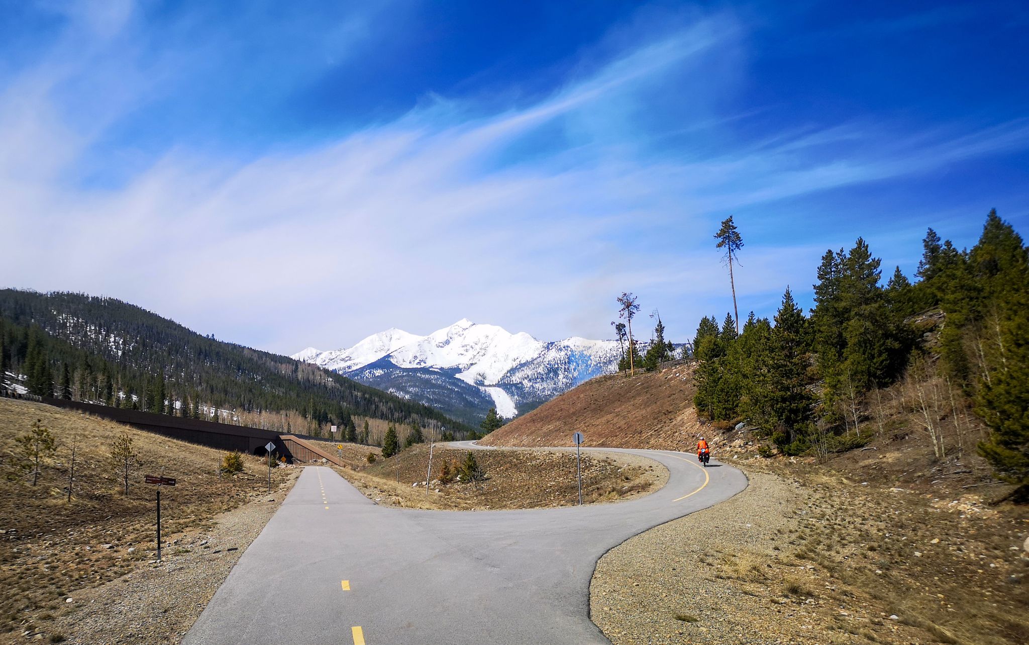 Cyclist on a paved path with forested hills and snow-capped mountains. - Lakeshore Perimeter and Crown Point Road Loop