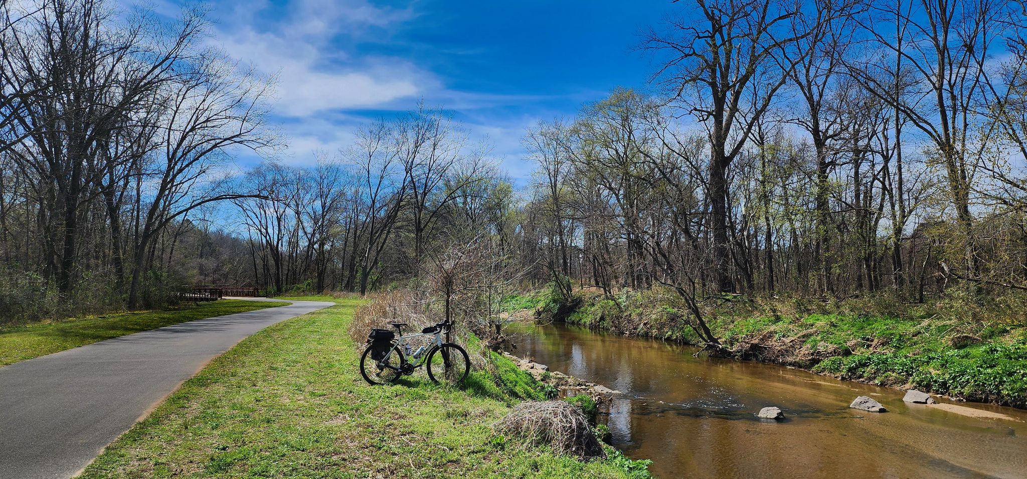 A bicycle parked on grass next to a paved path and a flowing creek. - Marsh Park loop from South Mecklenburg High School