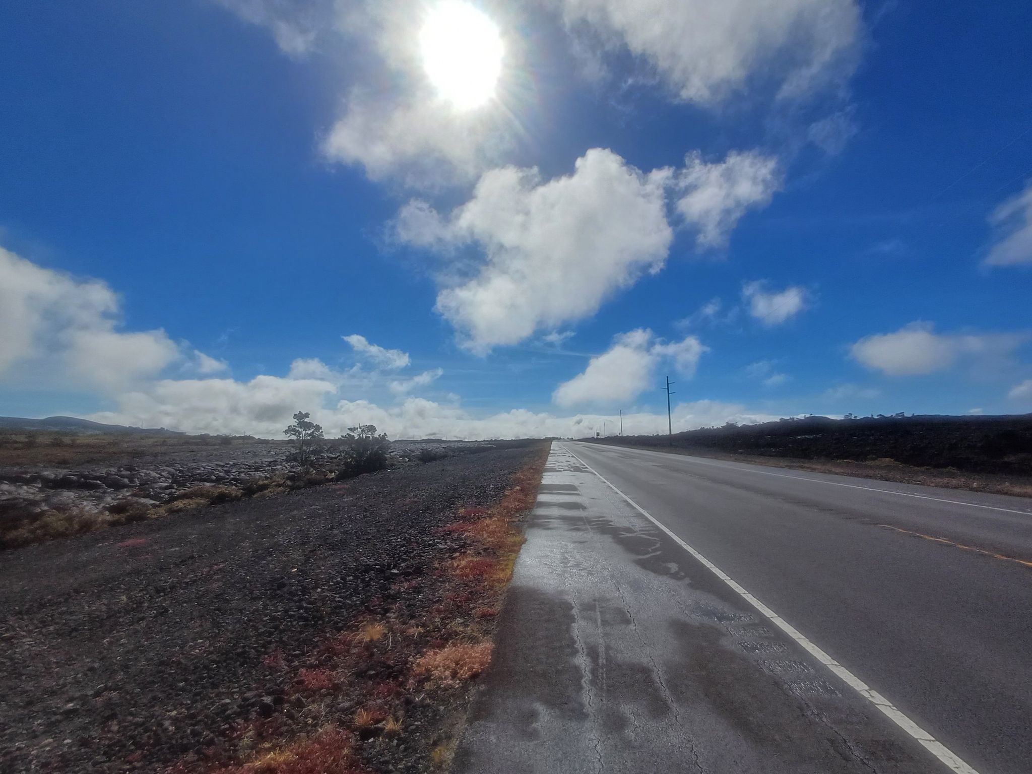 A long, empty road stretches through a barren landscape under a bright sun. - Mauna Kea Base via Hilo