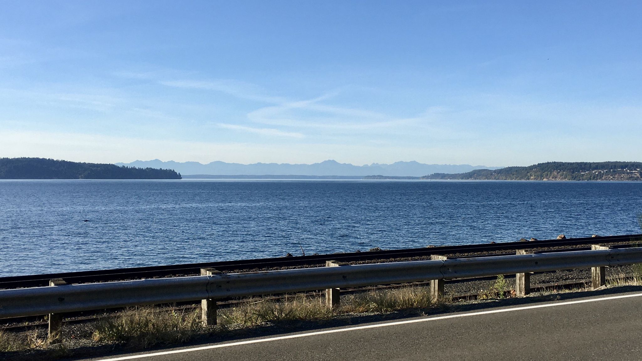 Sunny view of calm blue water, distant mountains, and a road with a guardrail. - Grandview Trail – Titlow Park loop from Tacoma Community College