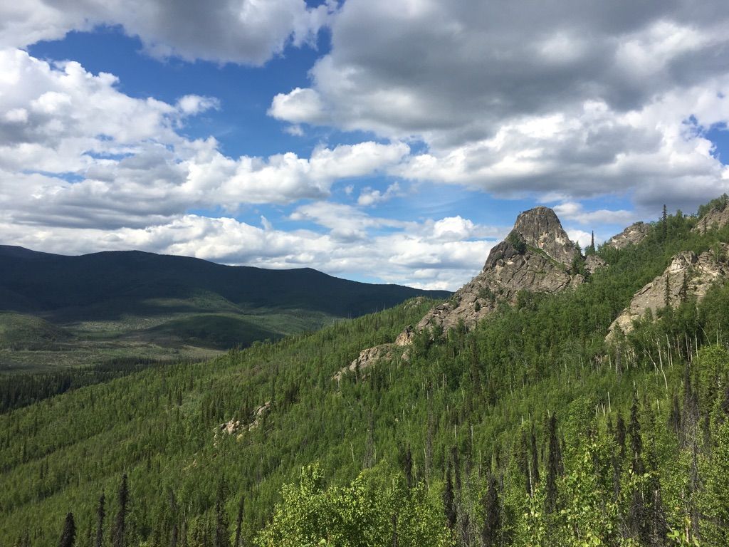 Rocky mountain peak surrounded by green forest under a cloudy blue sky. - Angel Rocks to Chena Hot Springs Trail
